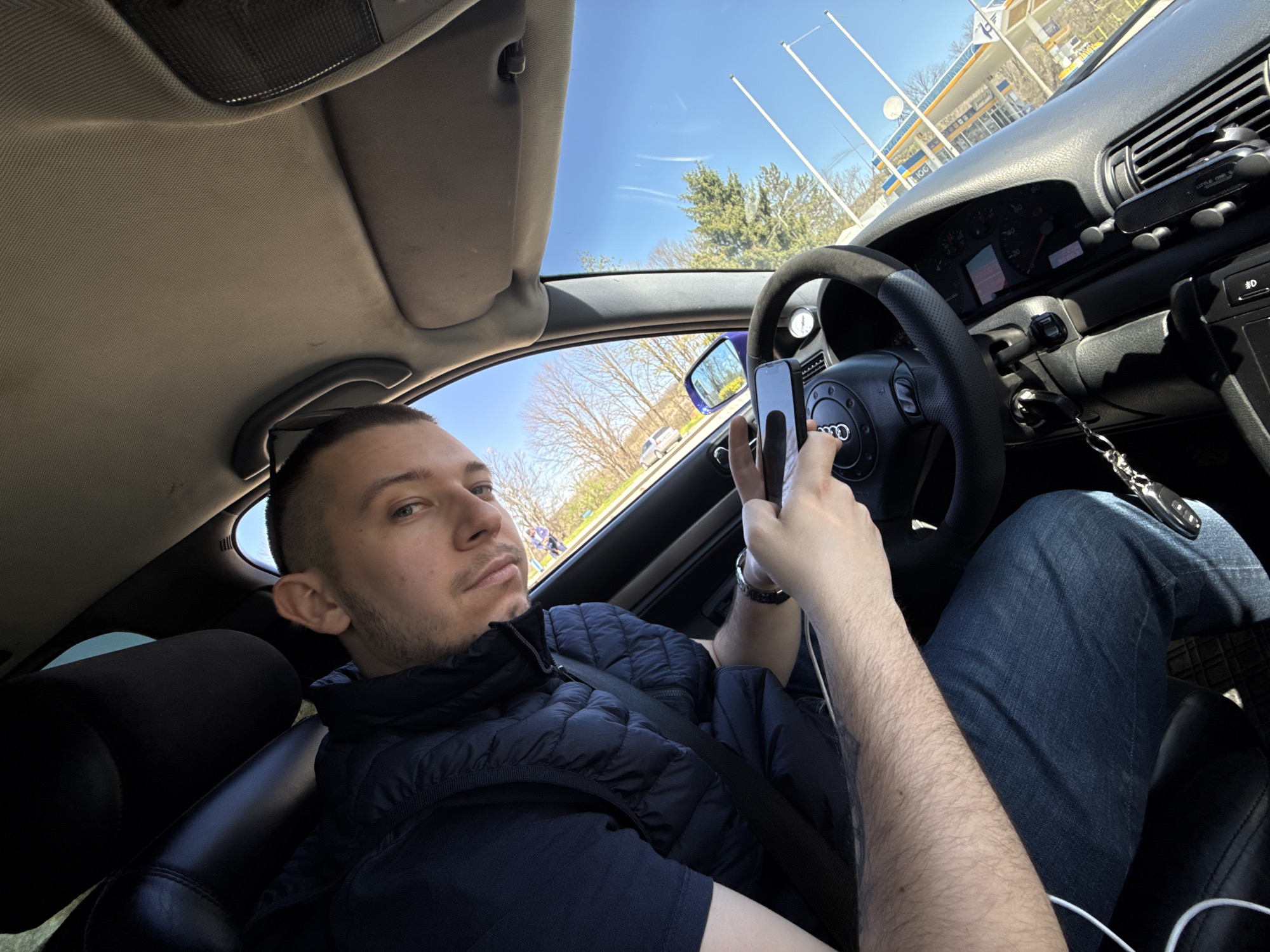 A man is in the driver's seat of an Audi. The car is stationary near a gas station, visible through the windshield. The man holds a phone in his left hand and appears to be taking a photo. He is wearing a dark puffy vest over a dark shirt, jeans, and a wristwatch. The car's interior is black leather. A key is dangling from the dashboard. The location appears to be Veliko Tarnovo, Bulgaria, due to the design of the gas station visible through the windshield.