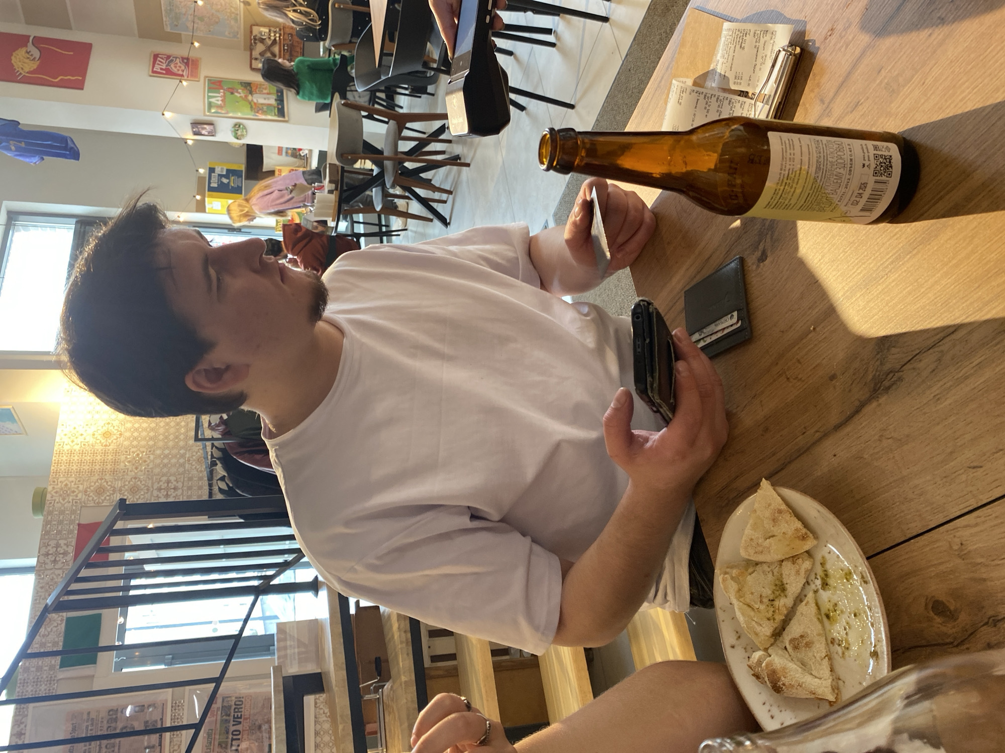 The image depicts a scene at a restaurant, likely in Izhevsk, Russia, judging by the Cyrillic text on the beer bottle. A man in a white shirt is seated at a wooden table. He is holding a smartphone in his right hand and a credit card in his left. On the table, there is a partially consumed plate of what appears to be pizza, a brown beer bottle, a wallet, and a receipt. The restaurant's interior is visible in the background, showing other patrons seated at tables, along with stairs and wall decorations. The atmosphere seems relaxed and casual.