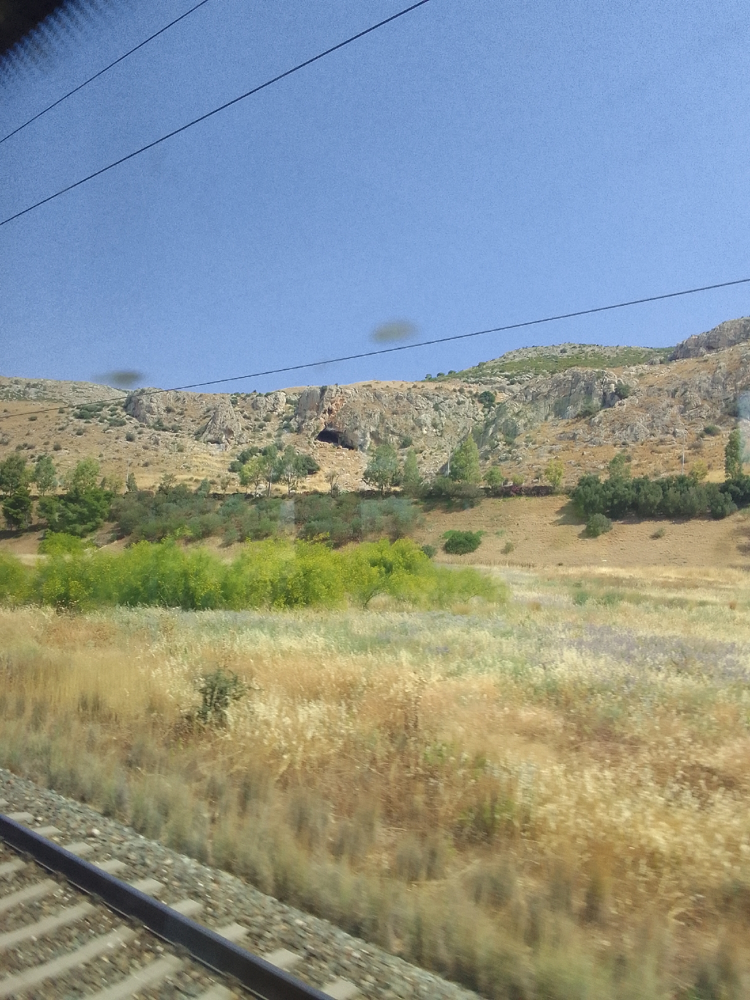 This media file depicts a rural landscape under a clear blue sky, suggesting daytime. The scene is viewed from a moving train, indicated by the blurred foreground of railway tracks. In the midground, a strip of vibrant green bushes stretches horizontally, contrasting with the dry, golden grasses and vegetation behind them. Beyond this, a hilly terrain rises, characterized by rocky outcrops and sparse vegetation. A notable feature on the hillside is a dark opening that appears to be a cave entrance. The overall setting suggests a Mediterranean climate, with the dry vegetation hinting at a warm, possibly summer season. There is no visible text in the image. The image captures a serene, natural environment, likely in the Municipal Unit of Fili, Greece.