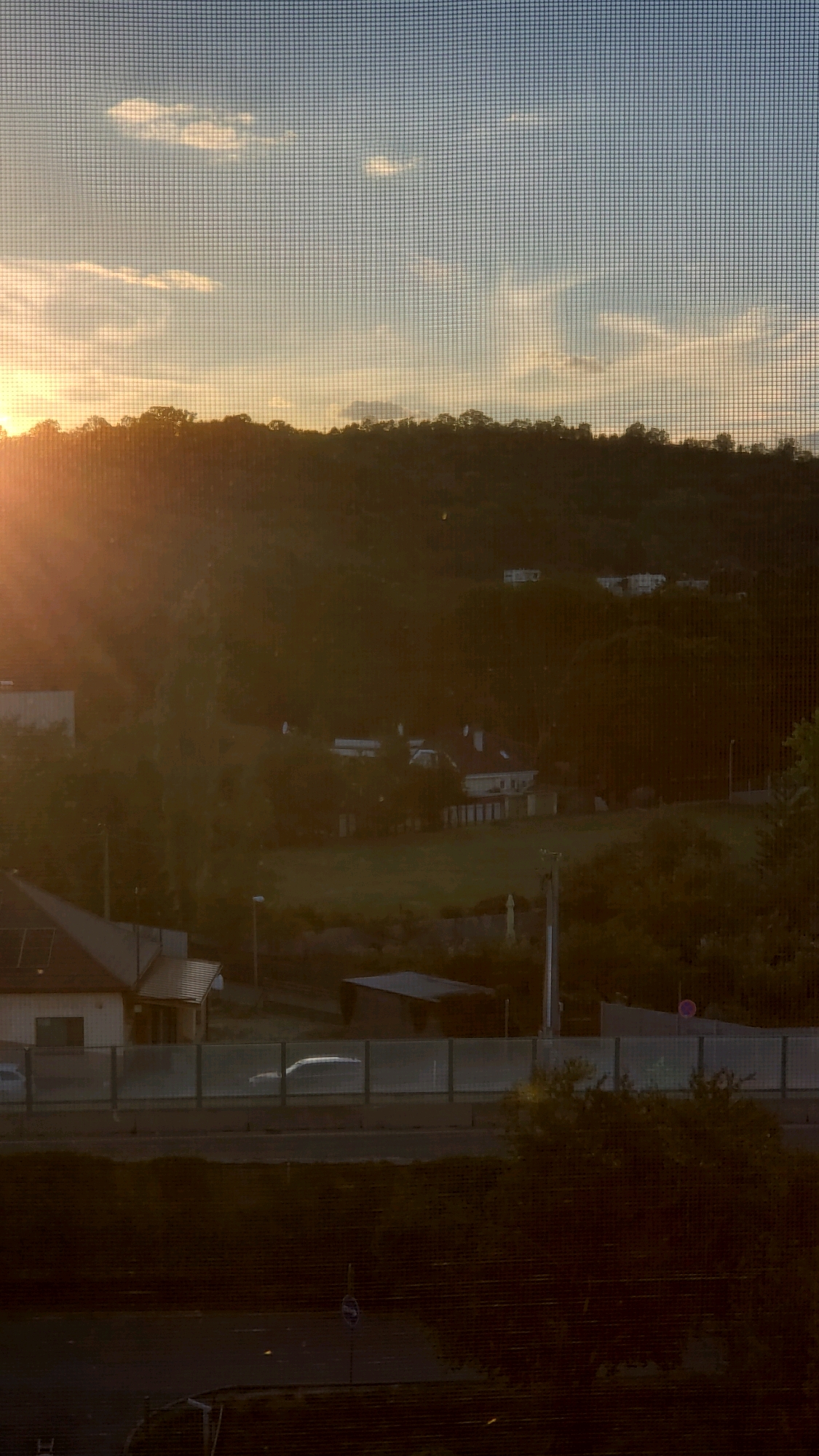 The image captures a late afternoon or early evening scene, likely from an elevated vantage point looking across a residential area towards a forested hill. The sun, low in the sky and casting long rays, is partially visible on the left side, illuminating the sky with warm hues of orange and yellow. Wispy clouds dot the pale blue sky.
In the mid-ground, a cluster of houses with red-tiled roofs are nestled amongst trees on the hillside. A well-maintained grassy area surrounds some of the properties, suggesting a suburban or semi-rural setting. A road or highway runs horizontally across the lower part of the mid-ground, with a noise barrier along its edge. A few vehicles are visible on this road, appearing as indistinct shapes due to the distance and lighting.
In the foreground, a dark, textured area suggests a wall or embankment, possibly made of brick. A street light is visible, and a blue traffic sign with a white symbol is mounted on a pole. The overall impression is one of a peaceful, somewhat hazy landscape transitioning from day to evening. The location is identified as Prešov, Slovakia.