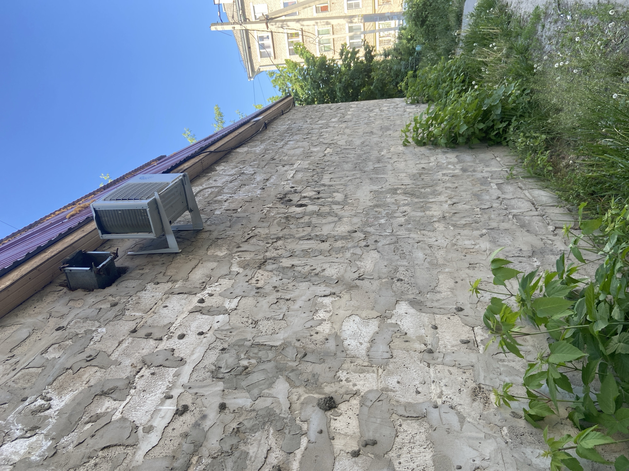 An upward-angle shot reveals a weathered, light-gray stucco wall with noticeable texture variations. At the top of the wall, green foliage and a portion of a multi-story building with yellow-toned walls and numerous windows are visible. On the left side of the wall, an air conditioning unit and its accompanying drain pipe are mounted. The sky is a clear blue. The image may be taken in Kazan, Russia.