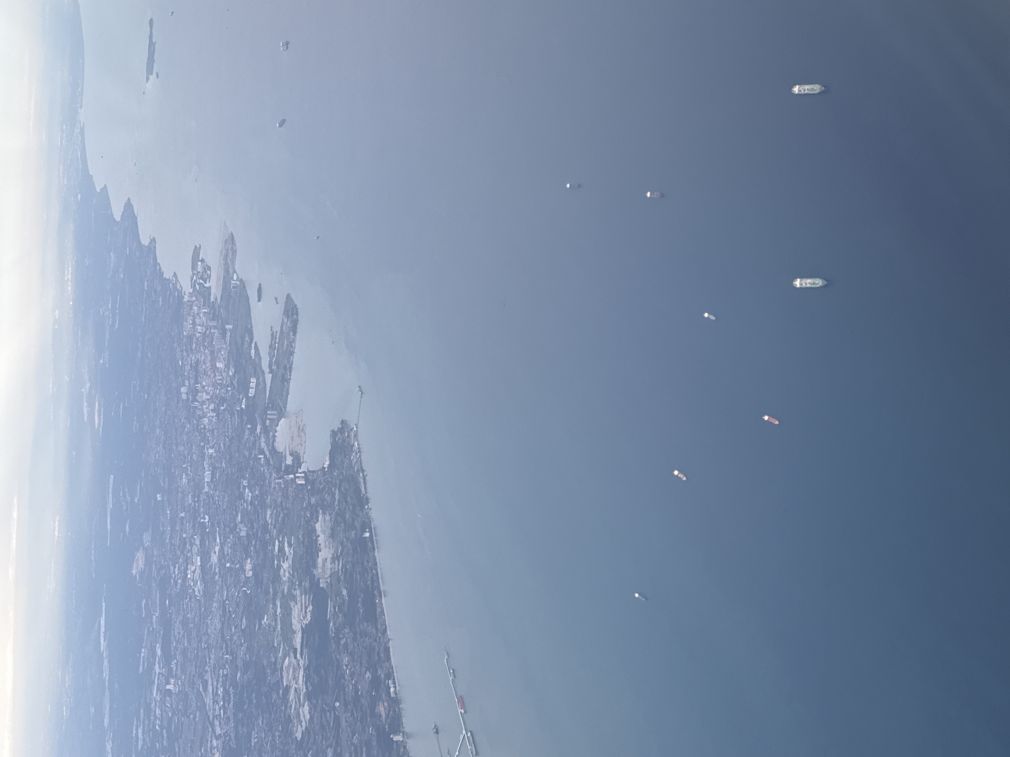 The image depicts an aerial view of the coastal area of Denpasar, Indonesia. A dense urban area is visible along the coastline, transitioning into water. Several ships are dispersed in the water, varying in size and distance from the shore. The environment is bright with a hazy atmosphere. There are no people visible in the image.