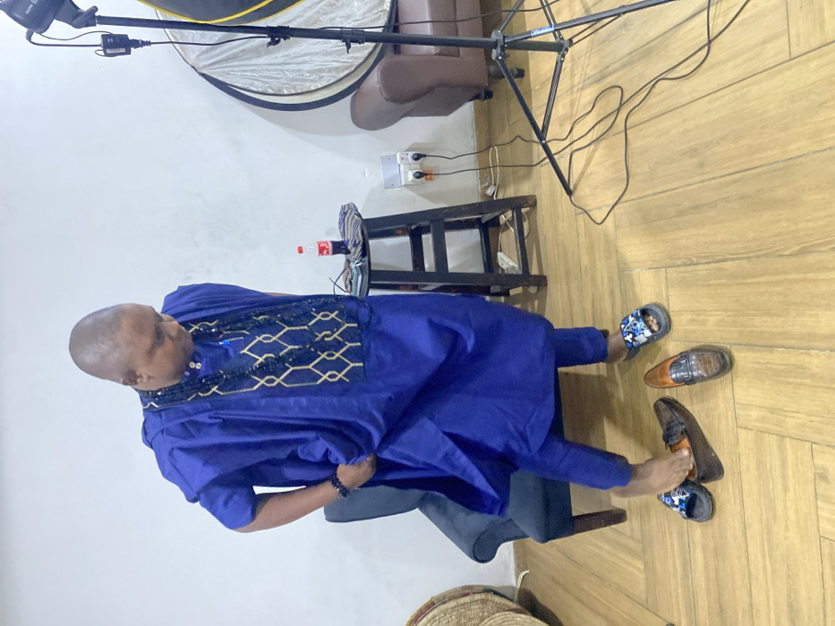 A man in a blue traditional African outfit stands on a wooden floor. He wears casual sandals, with a pair of dress shoes beside his feet. A black stool stands behind him, with a Coca-Cola bottle and a cell phone placed on top. A lighting rig is positioned in the background, along with a brown leather couch. A woven basket is partially visible in the lower left corner. The wall is white with a visible outlet.