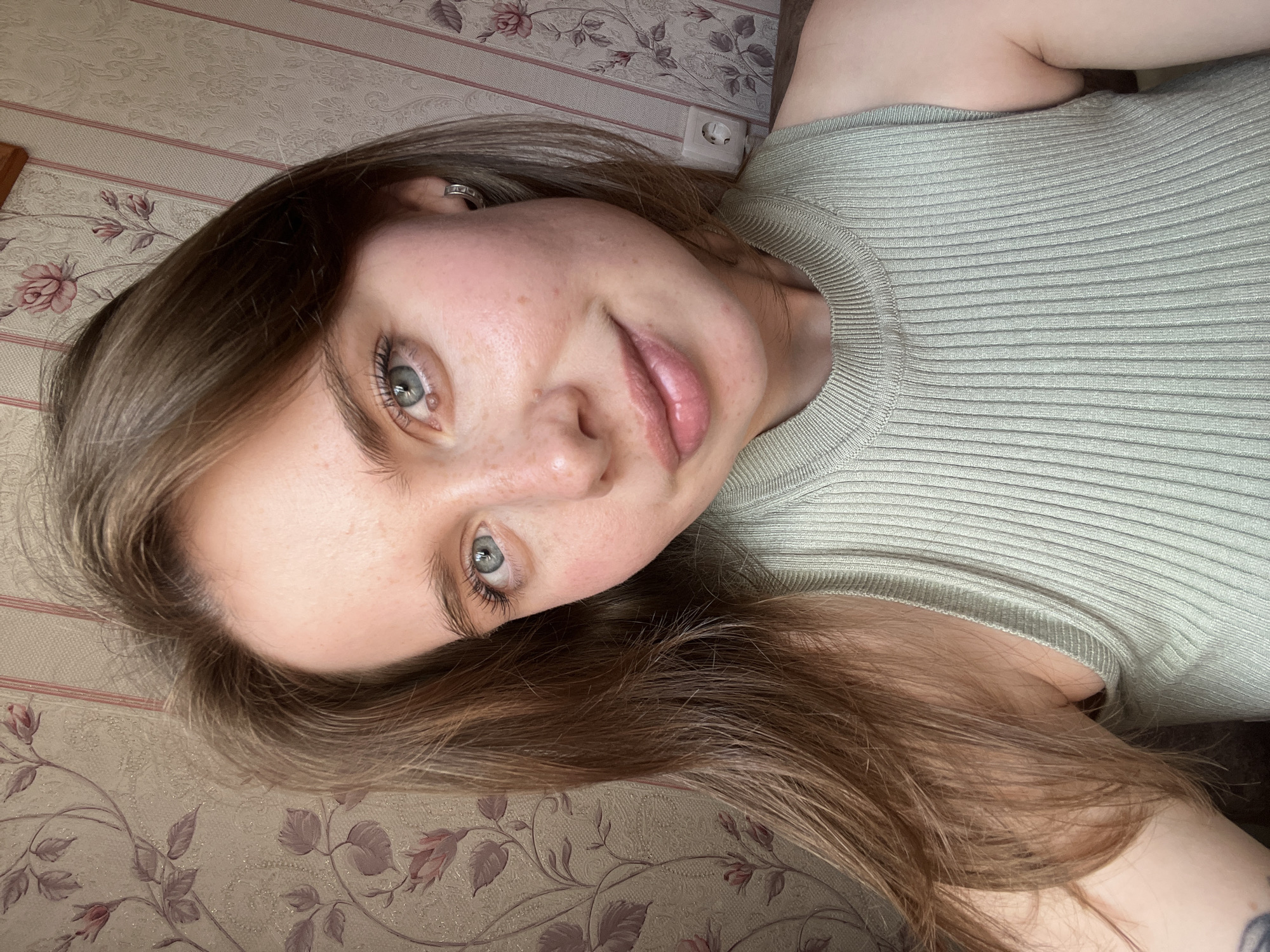 A close-up, selfie-style shot features a young woman with light brown hair and blue eyes looking towards the camera. She has fair skin with freckles and is wearing a light green ribbed tank top. The background consists of floral wallpaper with a pink and beige pattern. The image does not provide any specific information about the city of Chaikovsky, Russia.