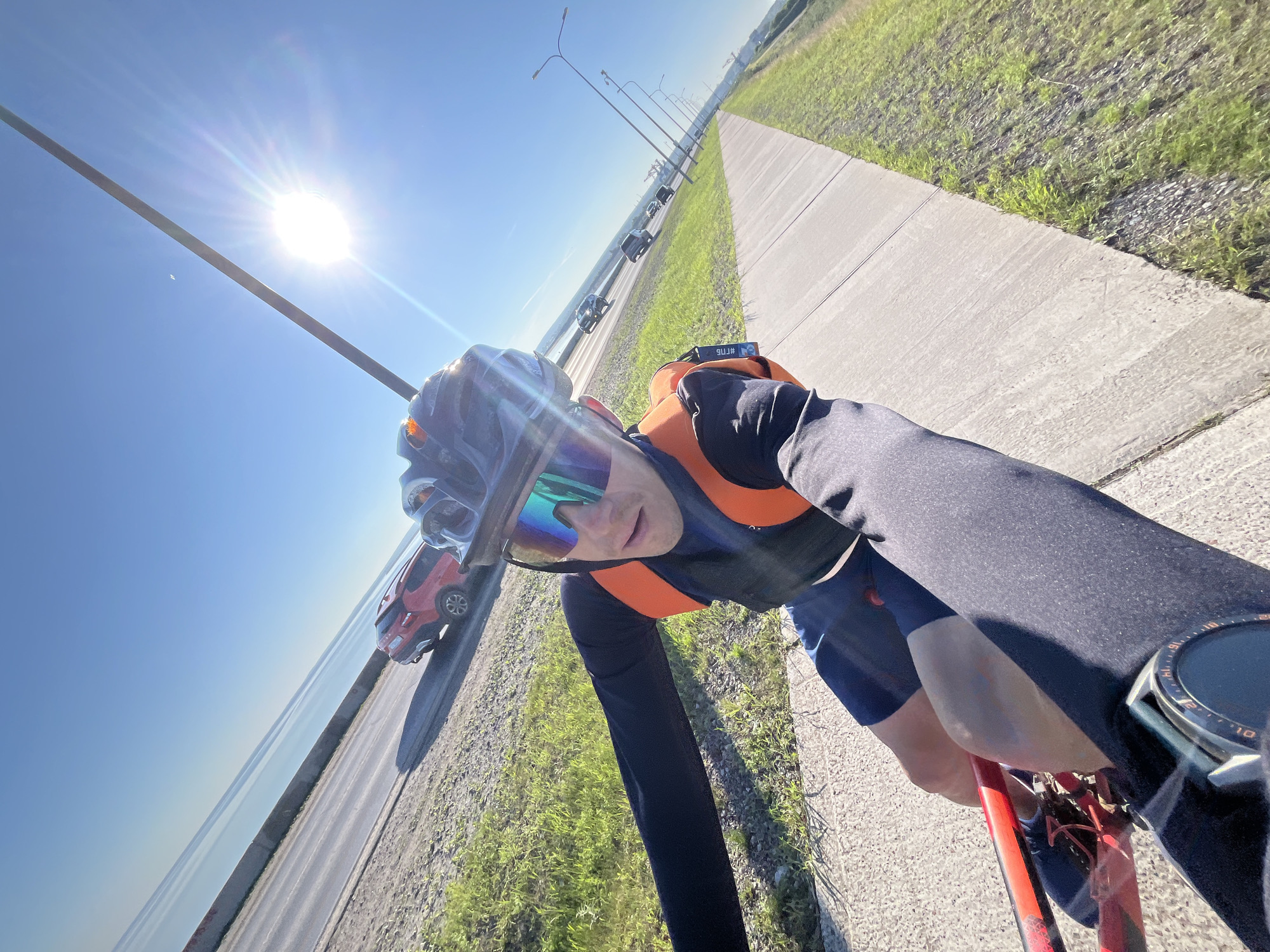 A first-person perspective shot shows a cyclist wearing a black long-sleeved shirt, blue shorts, a black helmet with a reflective visor, and an orange vest while riding a red bicycle. The cyclist is on a concrete path beside a road with several cars and a body of water in the background. The sun is bright in a clear blue sky. The scene appears to be taken in Chaikovsky, Russia, possibly near the Kama River, given the presence of a large body of water and industrial elements in the distance.