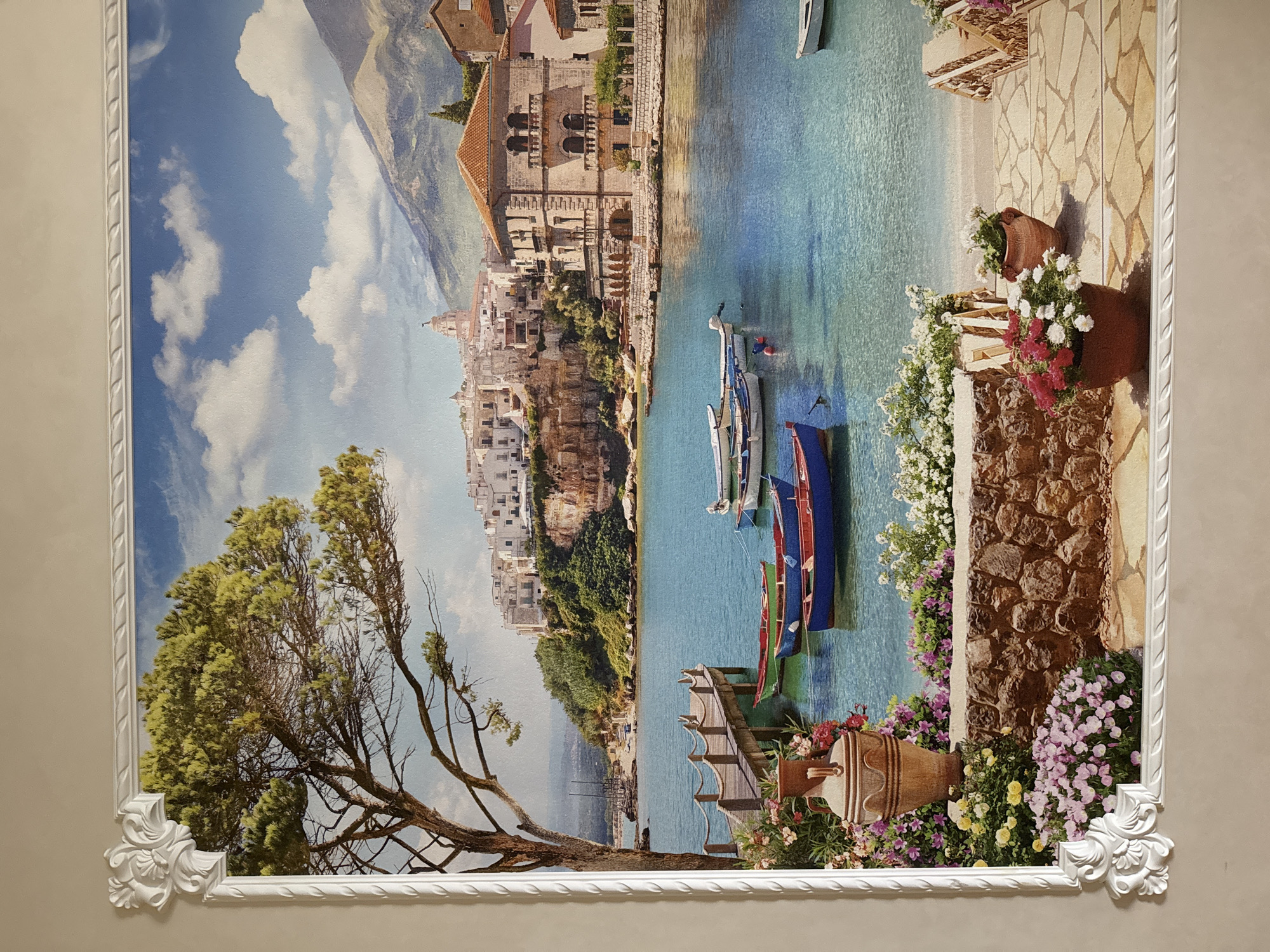 The image depicts a vibrant, picturesque scene of a coastal town. In the foreground, a large, ornate white frame surrounds the artwork. The artwork itself showcases a tranquil bay with several small boats floating on clear turquoise water. A wooden pier extends into the water from the left, adorned with a large ceramic vase and a profusion of colorful flowers. Behind the pier, a majestic tree with lush green foliage stands tall. Across the bay, a charming white-washed town is nestled on a cliffside, featuring buildings with red-tiled roofs and a prominent domed structure. Mountains rise in the background under a bright blue sky dotted with fluffy white clouds. The scene is detailed and realistic, creating a sense of idyllic serenity. There is no indication or depiction of the city of Sinyavino, Russia in this image.