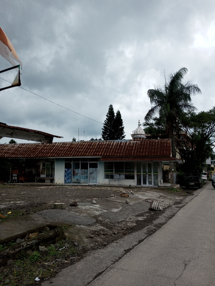 The image depicts an exterior street view in Melakudikadu, India, under an overcast sky with grey clouds. The foreground features an uneven, cracked paved and dirt lot with scattered rocks and debris, including a partially visible corrugated metal sheet. A small, overgrown drainage ditch runs along the bottom-left. To the right, a paved road extends diagonally into the background.
The central subject is a single-story building with a light-colored facade and a prominent, rusty, corrugated metal roof. The building features multiple large glass windows and doors across its front, some containing visible posters or promotional materials. On the far left, an older, partially deconstructed or damaged roof section extends, revealing an underlying structure.
In the mid-ground, a dark-colored utility truck is partially visible on the right side of the road, obscured by trees. Further down the road, a person in a light-colored top is partially visible near a motorbike or scooter. Overhead, thin wires traverse horizontally.
The background includes dense green foliage from various trees, notably a tall, dark green coniferous tree and a large palm tree on the right side of the frame. A white domed structure with a finial is visible above the building, positioned between the trees. In the top-left, a portion of a white and orange flag or banner is visible, attached to an unseen pole structure, with a triangular frame hanging below it. No explicit interactions are discernible among the visible subjects.