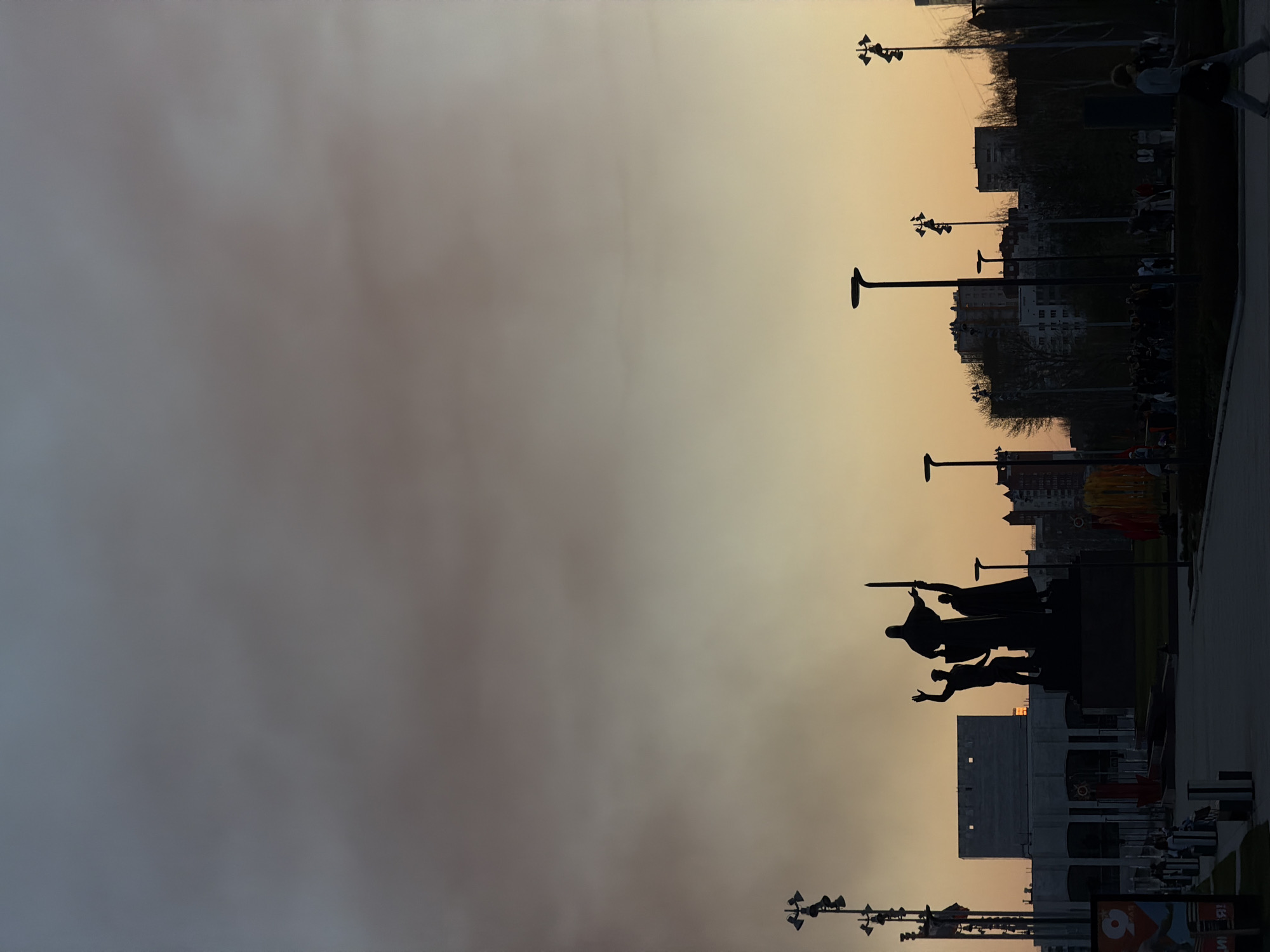 The image depicts a cityscape at dusk, with a prominent silhouette of a monument in the foreground. The monument appears to feature multiple figures, possibly a group of people in dynamic poses. Behind the monument, a large building is visible, also in silhouette. Streetlights line the area, and in the distance, several buildings, trees, and people are discernible. The sky transitions from a lighter shade near the horizon to a darker tone above. The image is rotated 90 degrees counterclockwise.
This scene is in Perm, Russia.