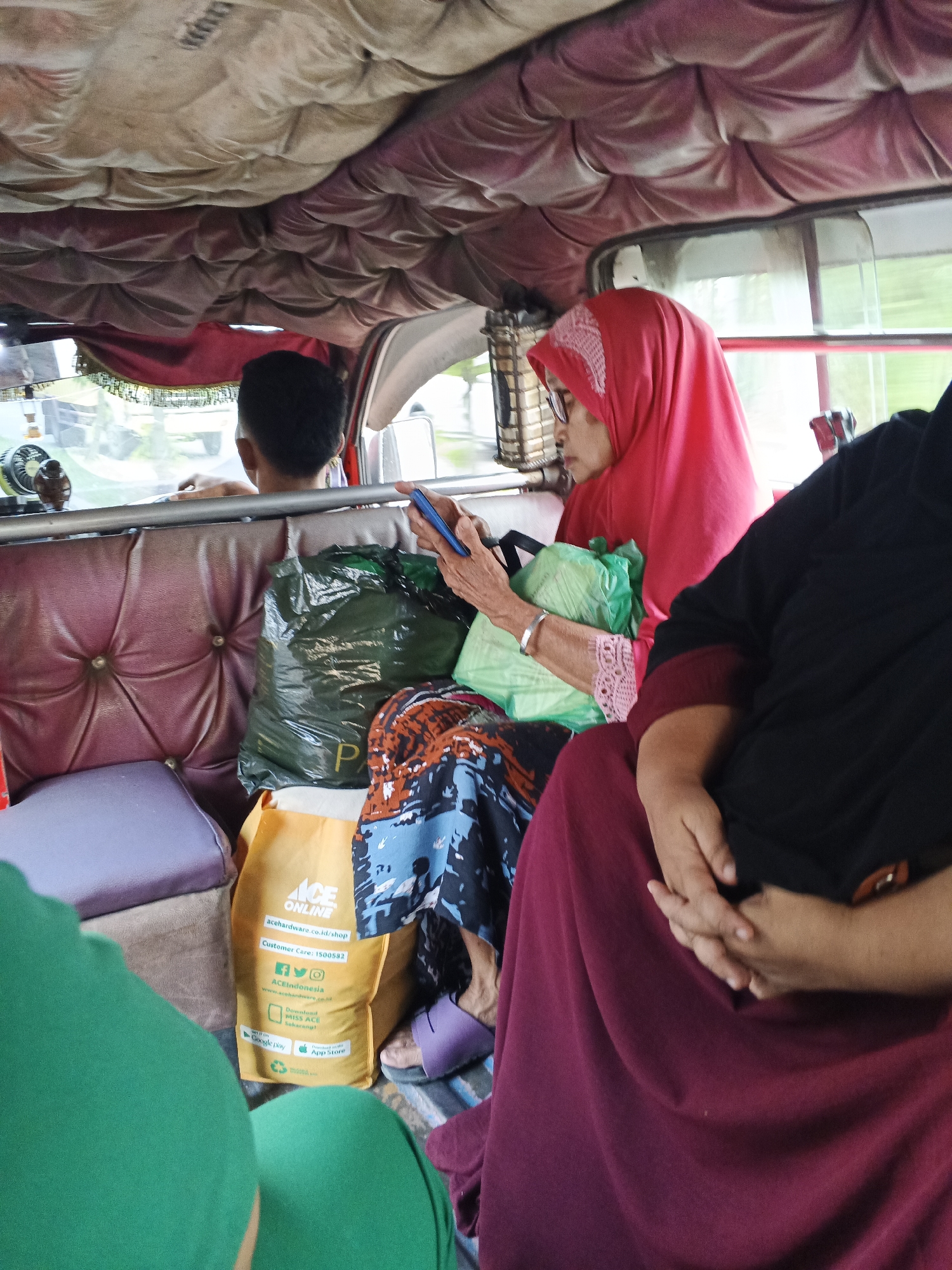 A woman wearing a red hijab and glasses is seated in the passenger area of a vehicle, possibly a van or a modified car, as indicated by the quilted upholstery and the interior ceiling. She is holding a blue smartphone in her hands and appears to be looking at it. Next to her, there is a green plastic bag and a yellow shopping bag with "ACE ONLINE" printed on it, suggesting a commercial area or recent shopping activity. Another individual, partially visible and wearing a black headscarf, is seated to her right. In the foreground, a person wearing a green shirt is also present. The background reveals the exterior through the vehicle's windows, showing a glimpse of a street or road, and trees, suggesting an urban or suburban environment. The overall scene is that of people traveling in a vehicle, possibly within the city of Tg Morawa A, Indonesia.