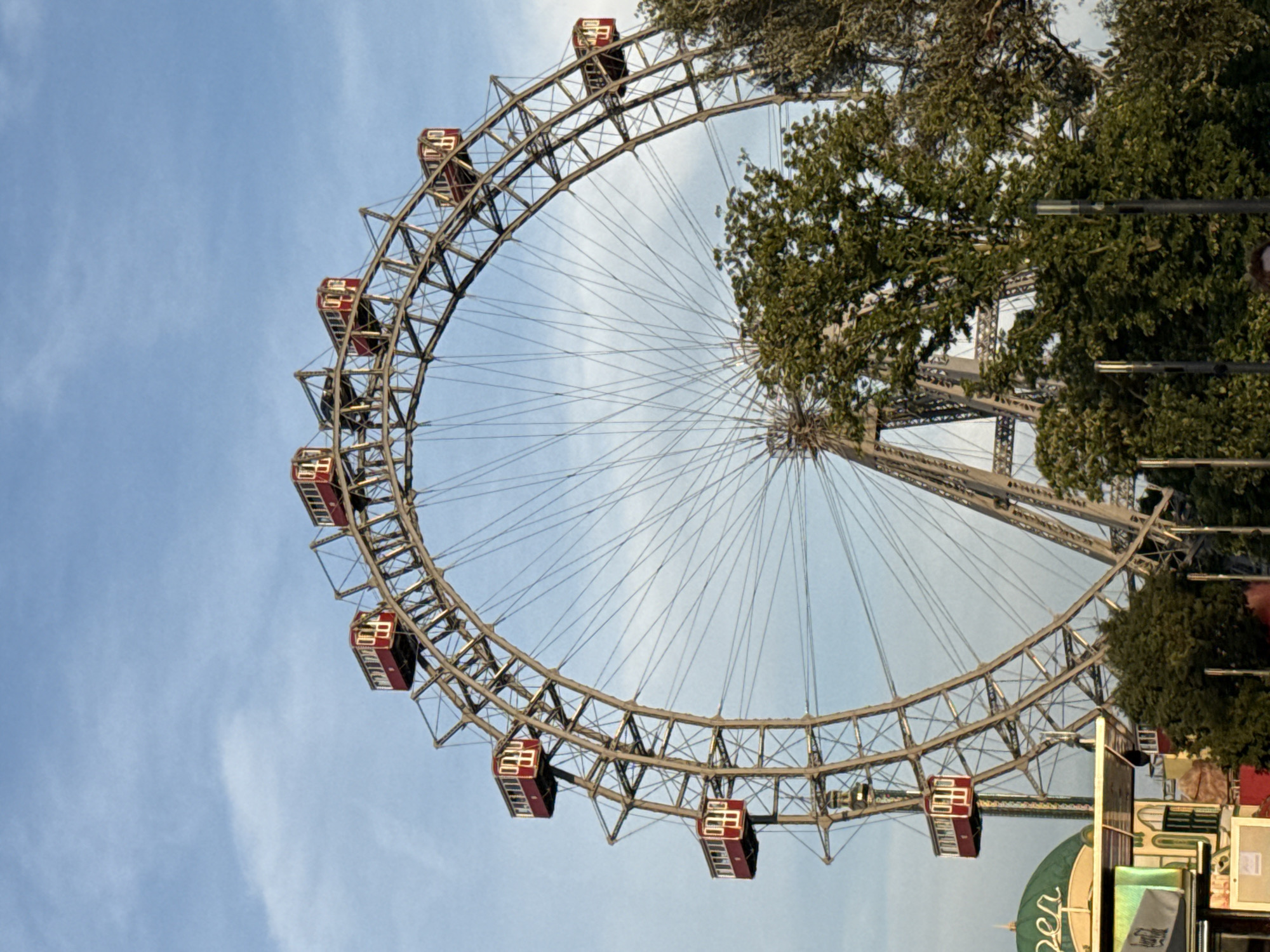 The image shows the Riesenrad, a historic Ferris wheel located in the Prater amusement park in Vienna, Austria. The Ferris wheel is a large, iconic structure with a weathered metal frame and several red gondolas suspended from its circular rim. It is captured from a slightly low angle, emphasizing its height and scale against a blue sky with scattered clouds. In the foreground and to the right, lush green trees partially obscure the view of the Ferris wheel's base and some of its central structure. To the left and in the lower part of the frame, parts of amusement park structures are visible, including a green domed roof and some colorful buildings. The overall scene suggests a pleasant day at an amusement park. There are no people clearly identifiable in the image, though it is possible a person's head is visible in the bottom right corner, out of focus.
