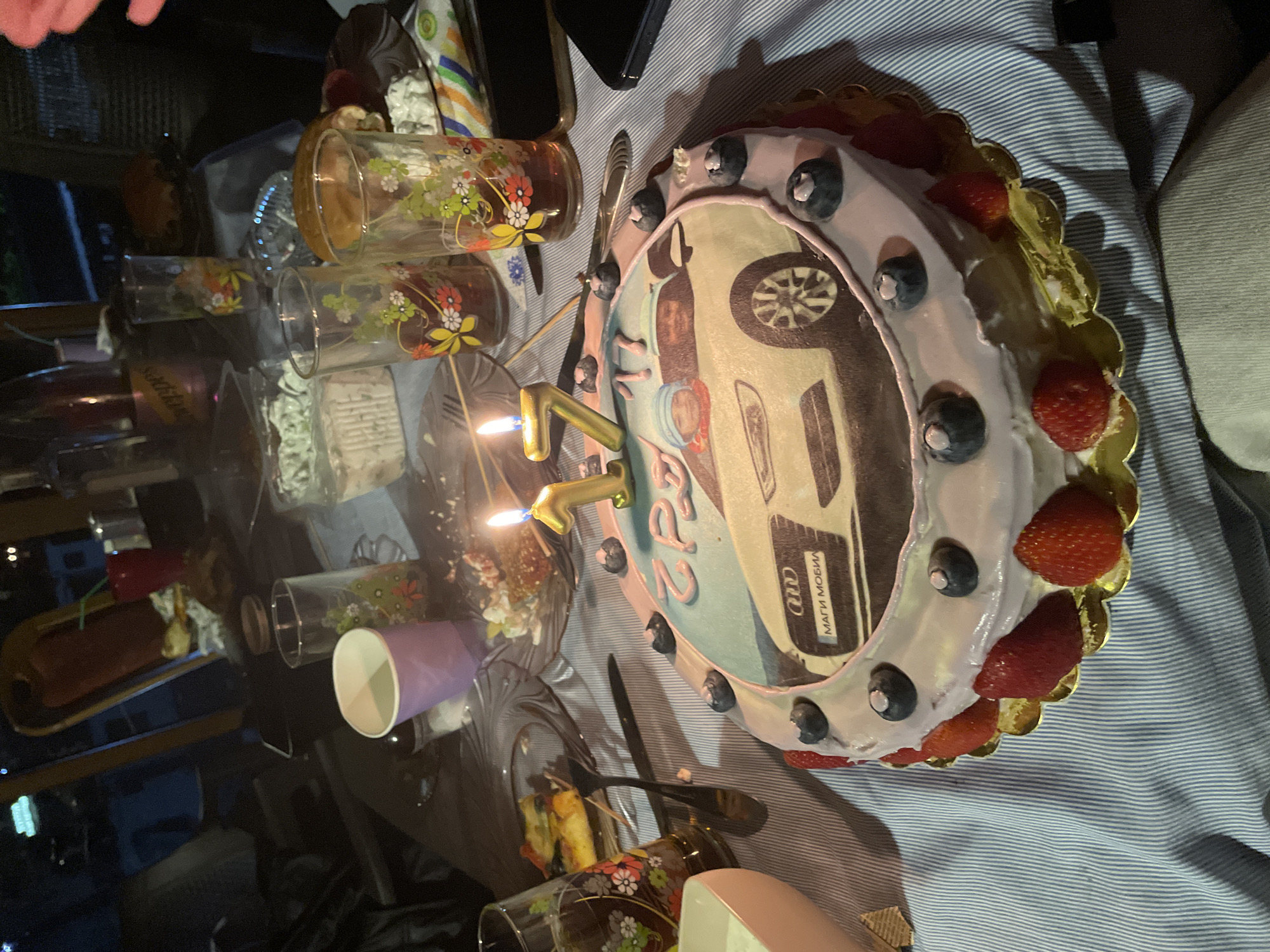 A brightly lit table holds a birthday cake with strawberries and blueberries, candles shaped like the number "22," glasses, plates with food, and a purple cup. The cake has an image of a car on it with the name "Audi" on it. The location is unknown, and there is no evidence of it being in Zlatograd, Bulgaria. The environment appears to be indoors. There are no people visible in the frame.