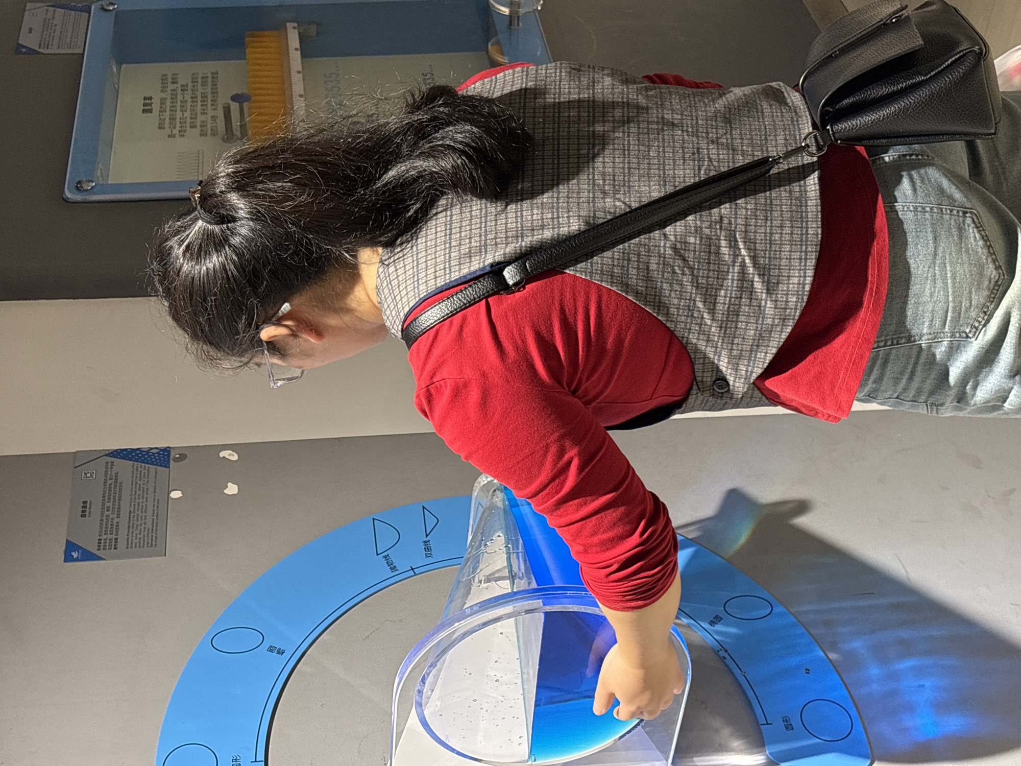 A person, likely a female, peers into a clear, cylindrical container containing blue liquid. The person is wearing glasses, a red top, a grey plaid vest, and blue jeans with a black purse slung over their shoulder. Their hair is pulled back in a low ponytail. Below the container is a light-blue diagram printed on the surface, indicating different geometric shapes. The scene appears to be inside an exhibit hall, evidenced by the educational displays on the walls. The city is Jiangjin, China.