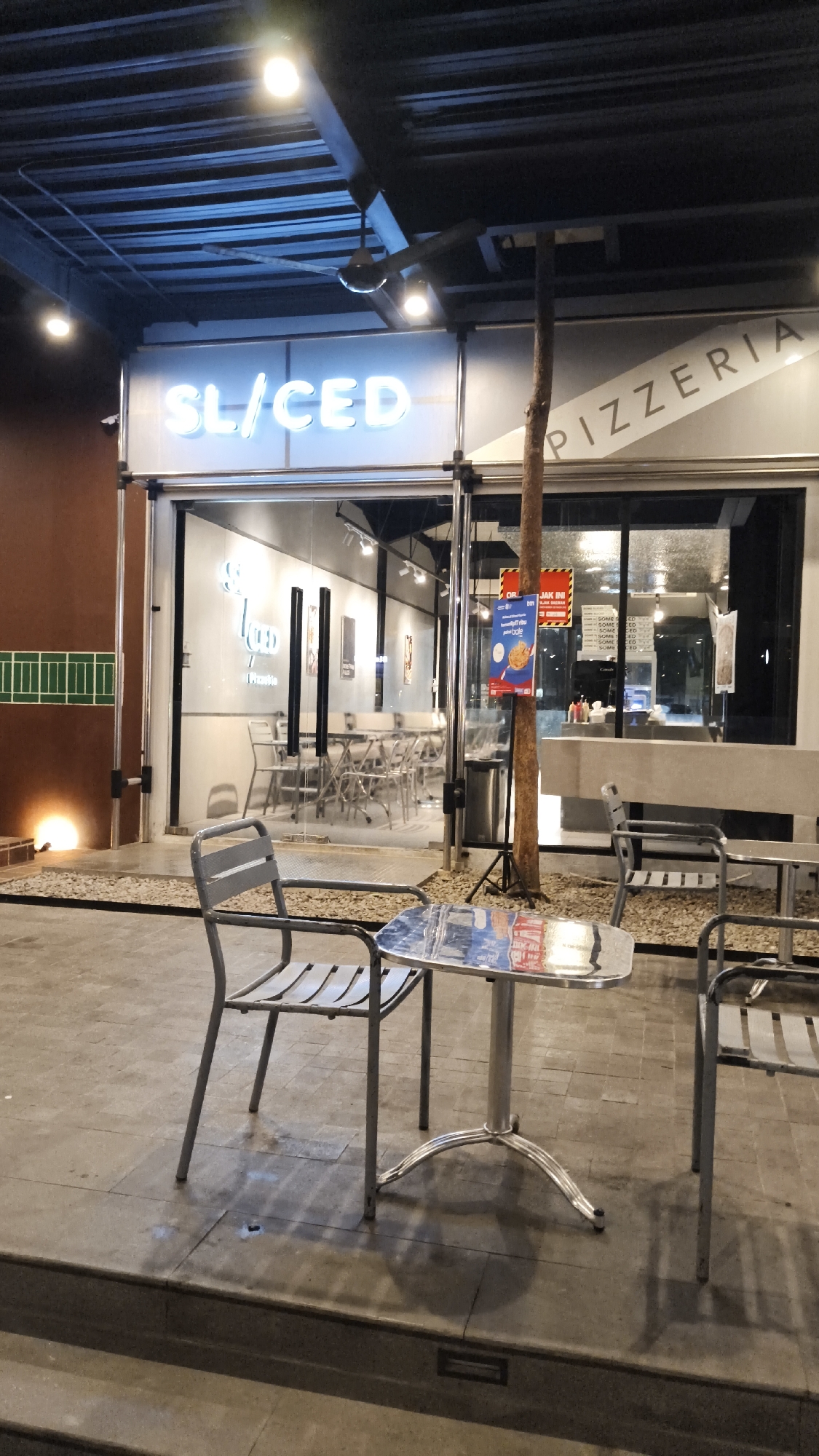 An exterior, medium shot captures a pizzeria establishment in South Jakarta, Indonesia, during nighttime. The pizzeria's facade features a prominent neon sign that reads "SL/CED" in glowing white letters, positioned above a glass-fronted entrance. To the right of the entrance, the word "PIZZERIA" is emblazoned diagonally on a light-colored surface. Inside the establishment, tables and chairs are visible, suggesting an indoor dining area.
In the foreground, a metal patio table with a reflective surface is set between two empty metal chairs. The chairs have slatted seats and backs, and their legs are angled. The ground surrounding the table and chairs consists of paved stone tiles and gravel. A tree trunk stands to the right of the entrance, its presence suggesting an outdoor element integrated into the pizzeria's design.
Several advertisements are affixed to the tree trunk and a nearby trash receptacle, adding to the urban aesthetic. The lighting appears to be a mix of ambient streetlights and interior illumination from the pizzeria, casting shadows on the ground. The overall scene conveys a casual, potentially evening dining atmosphere.