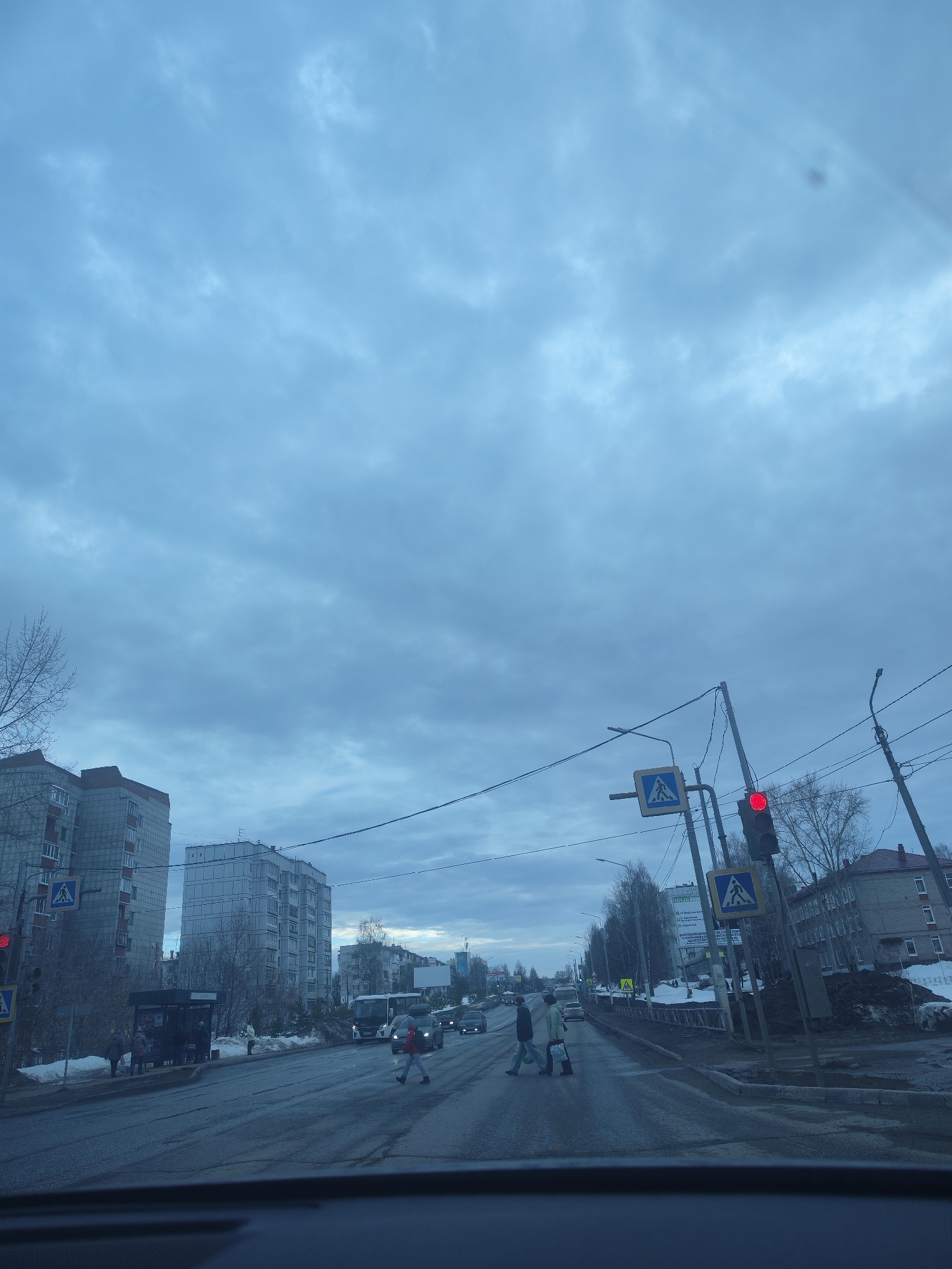 This image depicts a wide-angle view of an urban street intersection in Chaikovsky, Russia, during overcast daytime conditions, likely late winter or early spring. The perspective is from inside a vehicle, looking forward through a windshield, with the dashboard visible at the bottom of the frame.
The sky is uniformly grey and heavily clouded, dominating the upper two-thirds of the image. The ambient light is soft and diffused. Bare deciduous trees and bushes are visible on both sides of the street and along a median strip, indicating a non-growing season. Patches of snow and slush are present on the ground and accumulating along the roadside.
The asphalt road surface appears wet. A pedestrian crossing, marked by faded zebra stripes, is visible in the foreground. A traffic light, positioned above the right side of the road, displays a solid red signal. Accompanying the traffic light are two blue square signs with white triangles depicting a walking person, marking the pedestrian crossing. Several utility poles with overhead wires crisscross the frame on the right side.
On the road, three pedestrians are actively crossing from the left to the right side of the street, walking on the zebra stripes. The lead pedestrian is wearing a red jacket and dark pants. Behind them, another individual wears a dark jacket, and a third wears a light-colored jacket and dark pants, carrying a light bag. Three passenger vehicles are stopped in the right lane, facing away from the camera, presumably waiting at the red light. Further down the right side of the road, a white bus and another dark vehicle are visible.
On the left side of the street, a dark-colored bus stop shelter is present. Two additional individuals are near the bus stop: one standing outside near its entrance, dressed in dark clothing, and another partially obscured inside the shelter.
Architecturally, the left side of the street is dominated by two large, multi-story residential apartment buildings, typical of post-Soviet construction. The closest building has a grey facade with brown accents and a flat roof, while the one behind it is lighter grey. On the far right, further down the street, an older, multi-story building with a red-pitched roof is visible. A large, white rectangular sign, likely an advertisement or information board, is visible on the right side further down the road.
