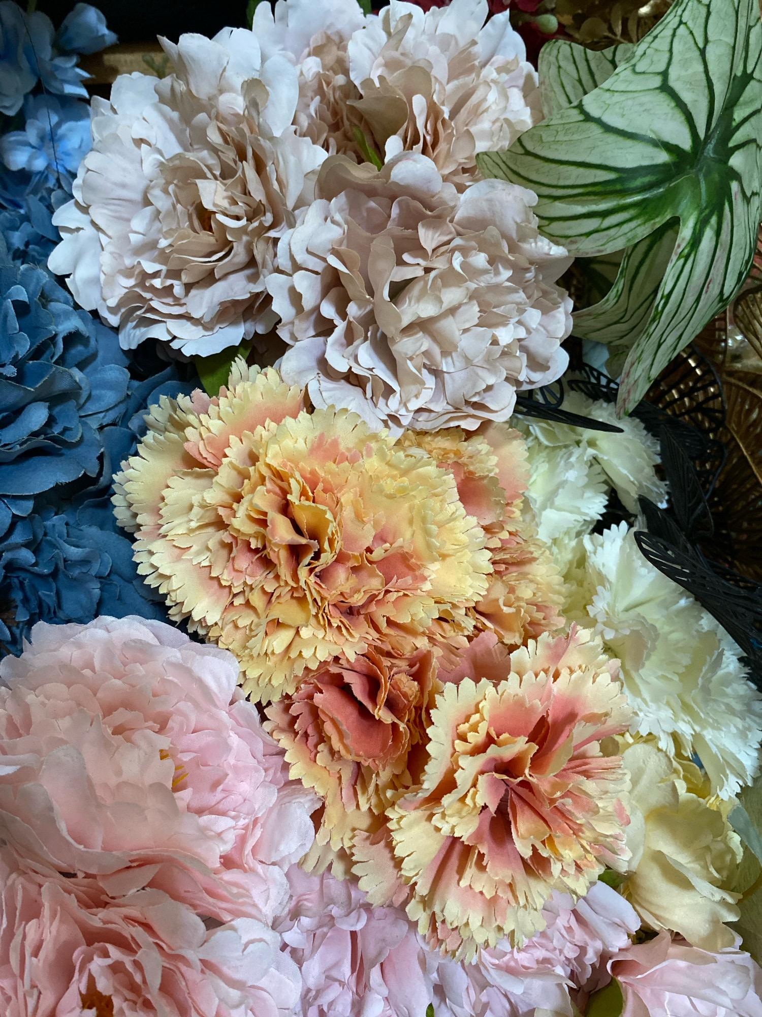 A close-up, overhead, medium shot captures a vibrant assortment of artificial flowers. The composition is dominated by large, layered petals in shades of pale pink, beige, and light yellow, interspersed with clusters of bright blue flowers and a striking green leaf with prominent white veins. The arrangement includes several carnation-like flowers with ruffled edges in peach and yellow hues, and other voluminous blooms resembling peonies in soft pink and beige. Some white flowers are also visible towards the right of the frame. The flowers appear to be part of a decorative arrangement, possibly a bouquet or a floral display. The background is dark and indistinct, focusing attention entirely on the textures and colors of the flowers. There is no visible indication of the city of Bandung, Indonesia, or any specific location within it.