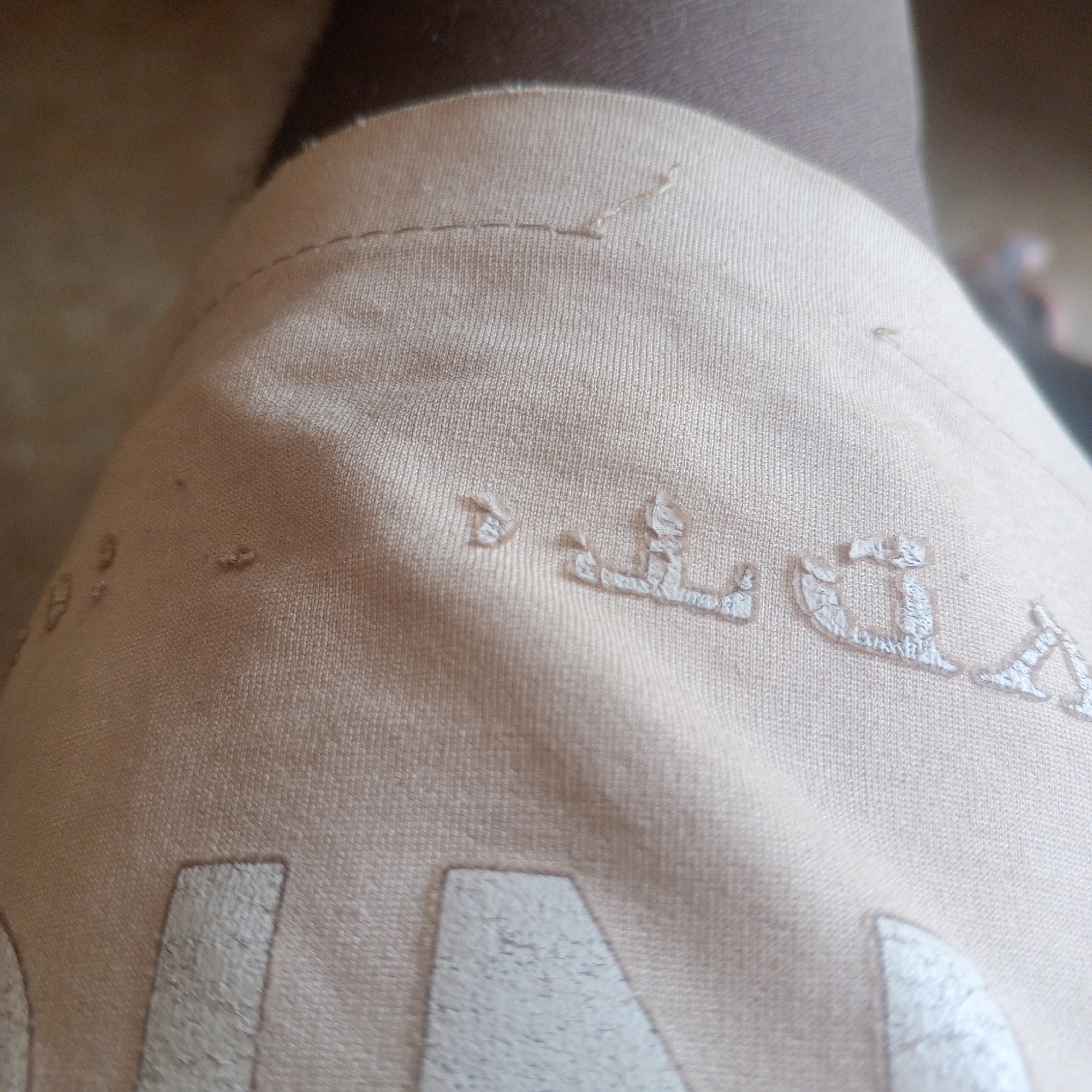 The image shows a close-up view of a light beige fabric, possibly clothing. There are some small holes or imperfections in the fabric. A partial inscription or logo is visible, appearing to be in a light color, possibly white or silver, and made of embroidered or printed letters. The letters are stylized and somewhat abstract. Some of the letters are clearer than others, but it is difficult to discern a complete word or phrase. The fabric appears to be a knit material. The background is blurred and shows a dark color, likely a person's leg or clothing. The image is taken from a close-up perspective, focusing on the texture and details of the fabric and inscription. There is no indication of the location in Zinna, Nigeria.