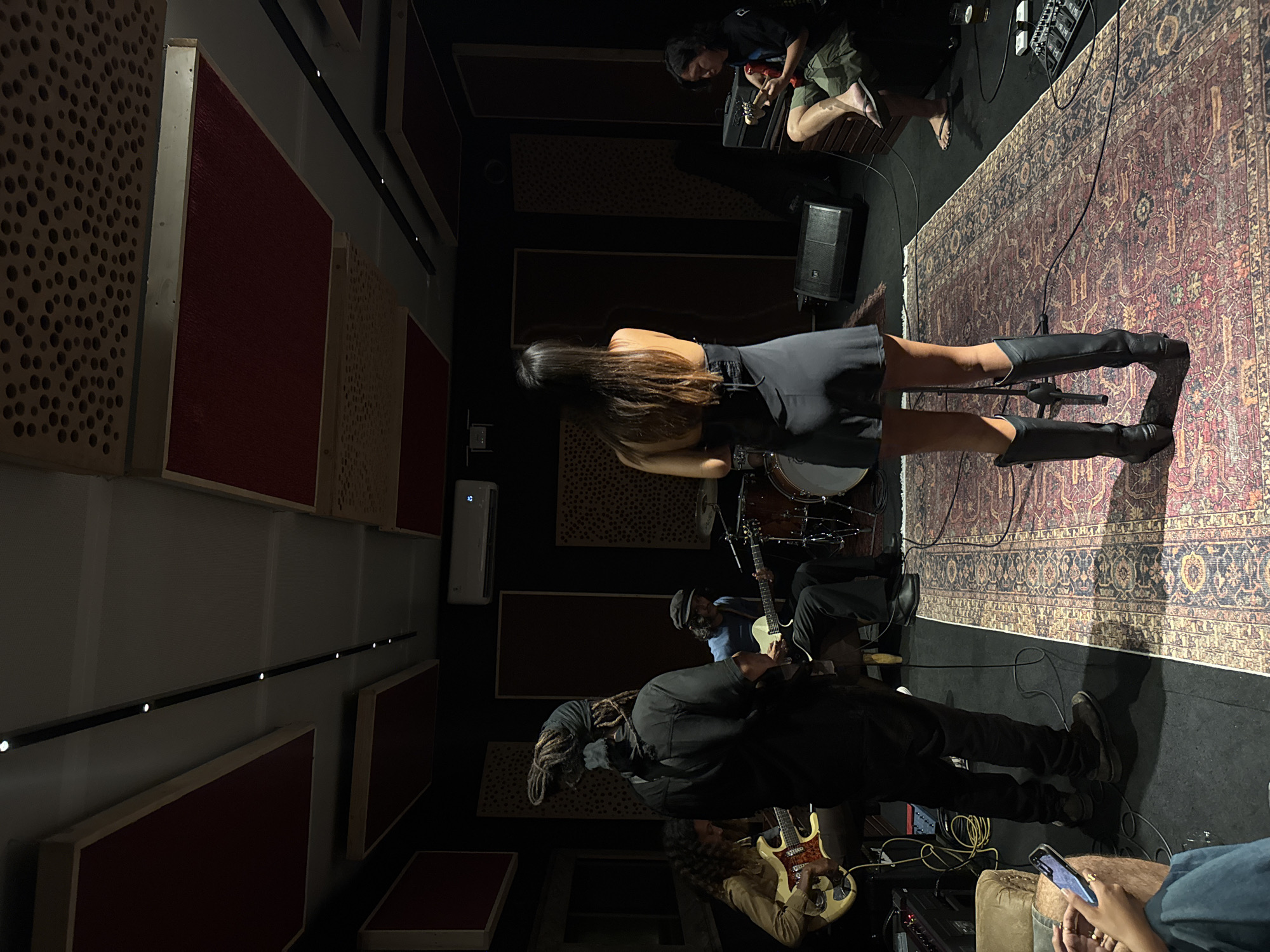 The image shows a band rehearsing in a music studio. The location is likely indoors, judging from the studio equipment and soundproofing panels on the walls.
People:
- A female drummer in a black dress and boots is visible from behind, playing the drums.
- Two guitarists are playing. One guitarist is in the foreground wearing all black, and another is slightly obscured in the background.
- A person is seated playing a guitar and looking at the camera in the upper right of the image.
- Another person is seated in the bottom right holding a mobile phone.
Subjects:
- Musical instruments (drums, guitars, amplifiers, effects pedals)
- A vintage-style rug on the floor.
Objects:
- Acoustic panels on the walls
- A sound system speaker
- Electrical cables
- An air conditioning unit
Actions:
- Band members playing musical instruments
Interactions:
- Musicians performing together as a band
Environment:
- A music studio with soundproofing, recording equipment, and musical instruments
The location in North Kuta, Indonesia cannot be verified solely based on the visual information in the image.