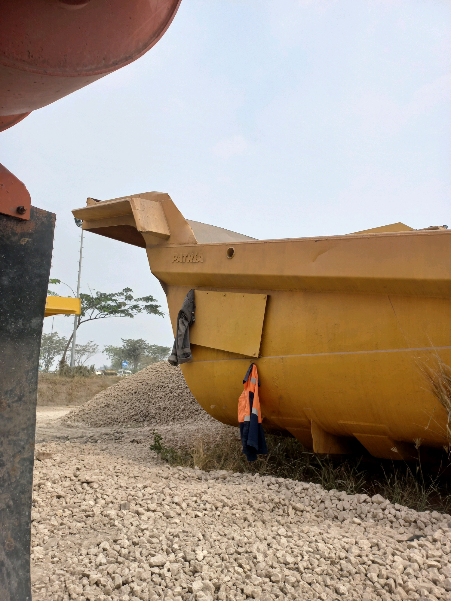 The image depicts an outdoor construction or industrial scene in Paringin, Indonesia, characterized by large yellow construction equipment and piles of gravel. In the foreground, a close-up view shows the lower portion of a massive yellow dump truck bed, angled upwards and to the right. A dark grey jacket is hung on the side of the truck bed, and a high-visibility vest, orange and navy blue, is hanging below it. The ground is covered in light-colored gravel, with some dried grass and vegetation visible. In the background, a large pile of gravel is visible, and beyond that, trees and a utility pole are present. The sky is overcast and light blue. The overall impression is one of an active work site, possibly a quarry or a material handling facility.