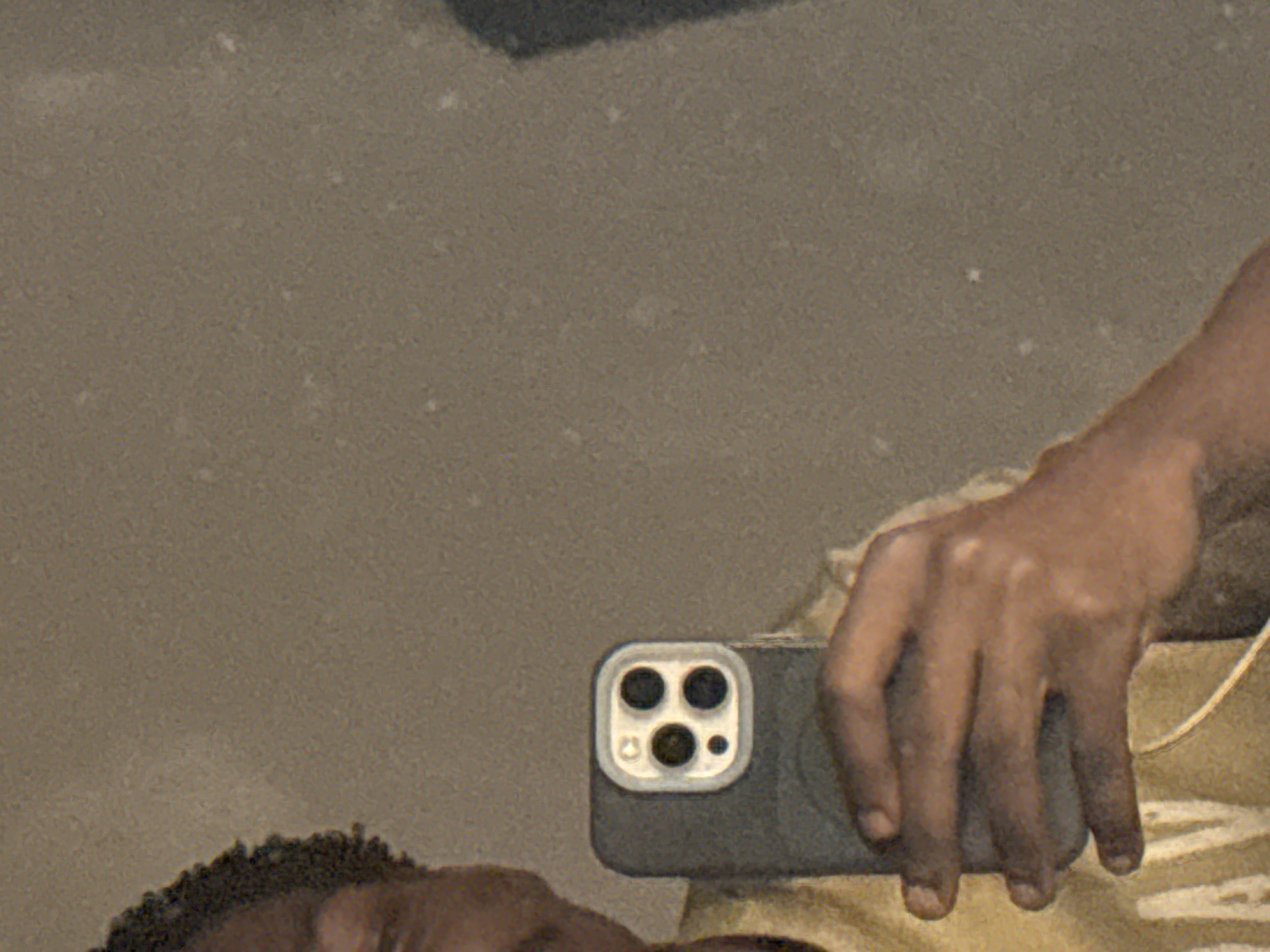 A close-up, waist-up selfie taken in Uyo, Nigeria, shows a person holding a smartphone, likely in a residential setting given the plain wall background. The individual, with short, dark, curly hair, is wearing a yellow t-shirt with what appears to be lettering, partially visible. The smartphone, held in their right hand, has a distinctive camera module with four circular lenses arranged in a square pattern, set within a silver bezel. The wall behind the person is a neutral, speckled gray, suggesting an indoor environment. The lighting is dim, and the image has a grainy texture, possibly due to low light conditions or the camera's settings. The overall impression is casual and intimate, a snapshot of everyday life captured in a personal moment.