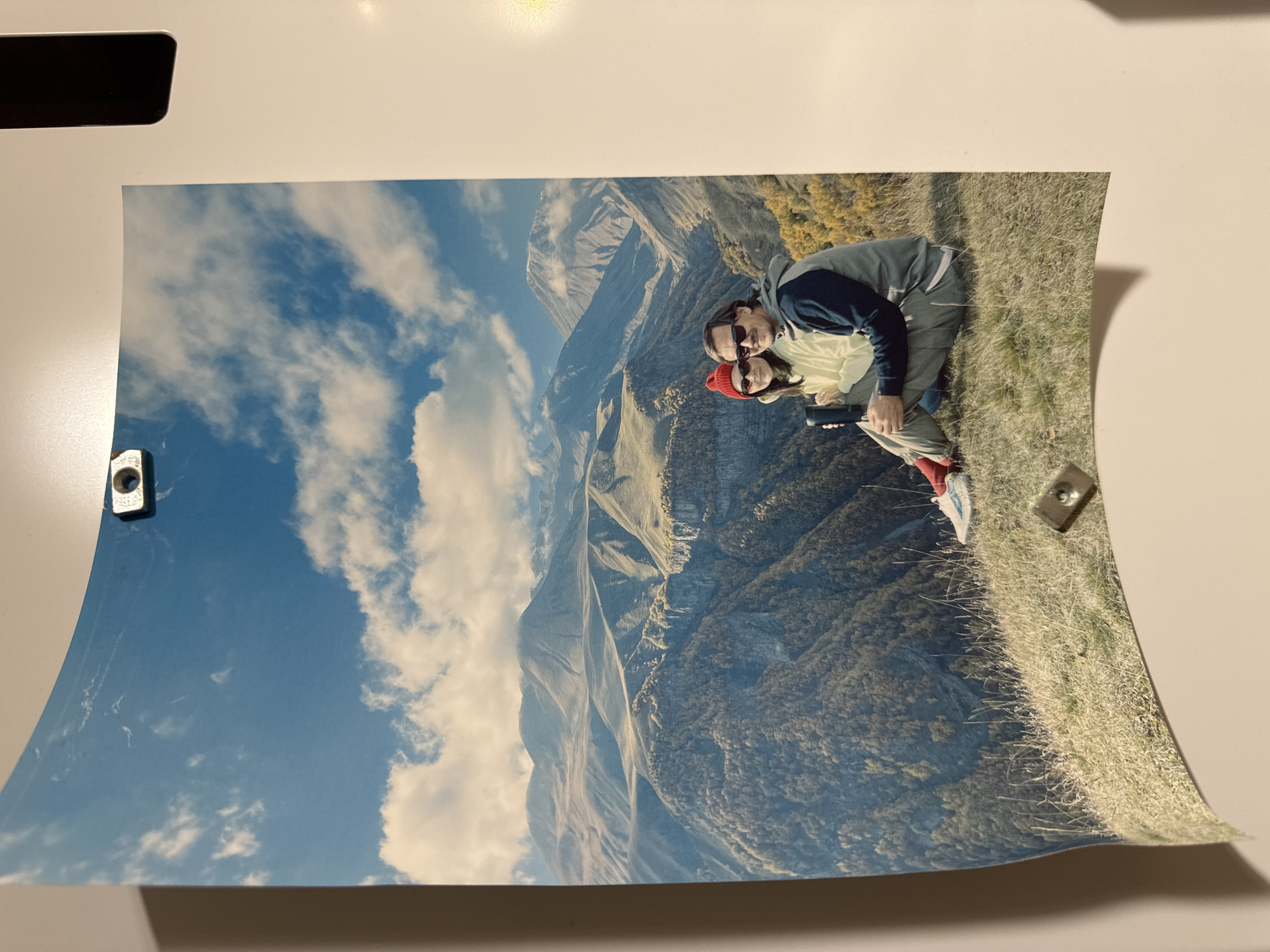 A photograph of a man and woman standing on a grassy hillside with a mountain range and blue sky with clouds in the background is held to a white surface by two small, silver-colored, square magnets with holes. The man is wearing sunglasses and a jacket. The woman is wearing a red hat and sunglasses. They are holding a small black object. The photograph is curled. There is a black object in the upper left corner of the surface holding the photograph. The location is not Izhevsk, Russia.