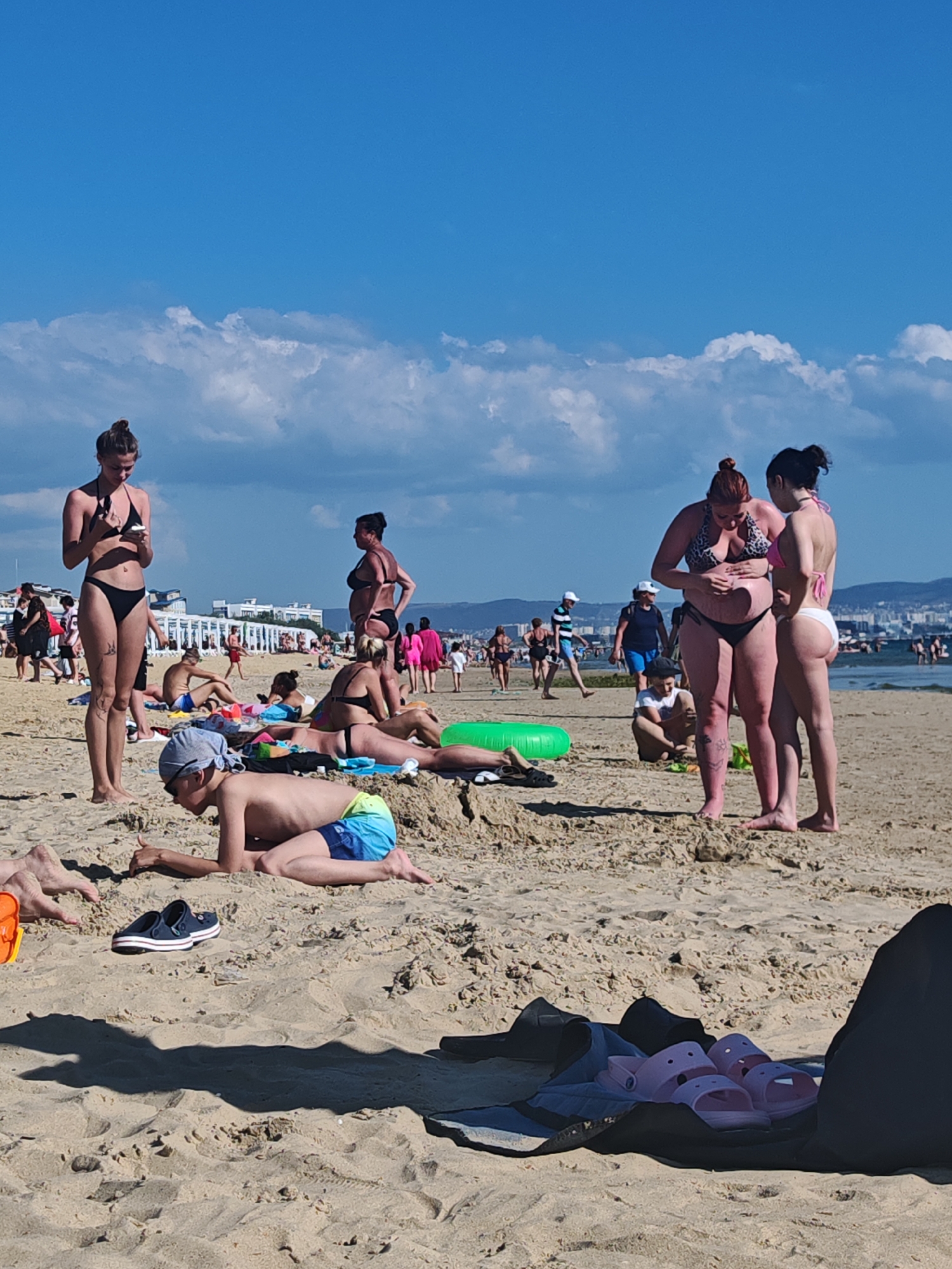 This media file captures a sunny day at a sandy beach, likely in Anapa, Russia. Numerous people are present, engaged in typical beach activities. Several individuals are sunbathing on towels, while others are standing and conversing. A young boy in the foreground is lying on his stomach, appearing to be playing or resting on the sand. He is wearing blue and green swim trunks and a grey bandana. A pair of dark blue sandals with white trim is visible in the sand near him. In the background, more people are scattered across the beach, with some near the water's edge and others further back. Buildings and a cityscape are visible in the far distance, suggesting the beach is situated near a populated area. The sky is bright blue with scattered white clouds, indicating pleasant weather. The overall atmosphere appears relaxed and leisurely, typical of a summer day at the seaside. There is no visible text in the image.