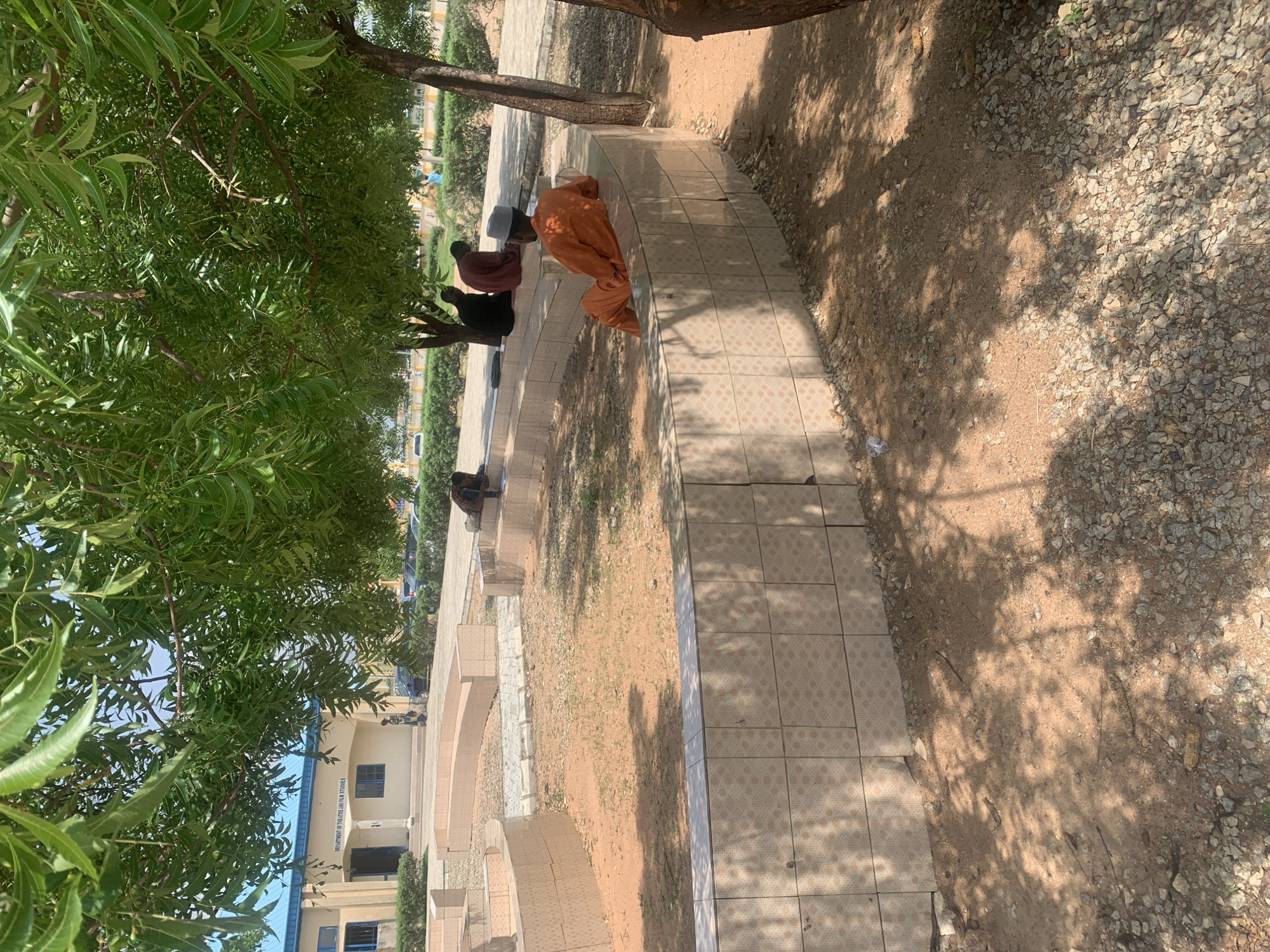 In Jalingo, Nigeria, in front of the Department of Theatre and Film Studies building, four individuals are seated outdoors under the shade of trees. The foreground features a tiled, circular seating area with an earthy floor. One person in an orange robe and a grey cap is seated on the edge of this area, facing away from the camera, looking towards the building. Further back, three other individuals are seated on a similar tiered, tiled structure. One person in a black robe and two in maroon robes are present. The building behind them has a blue roof and beige walls with visible windows and an entrance. The environment is characterized by sparse vegetation, gravel, and shadows cast by the overhead foliage.