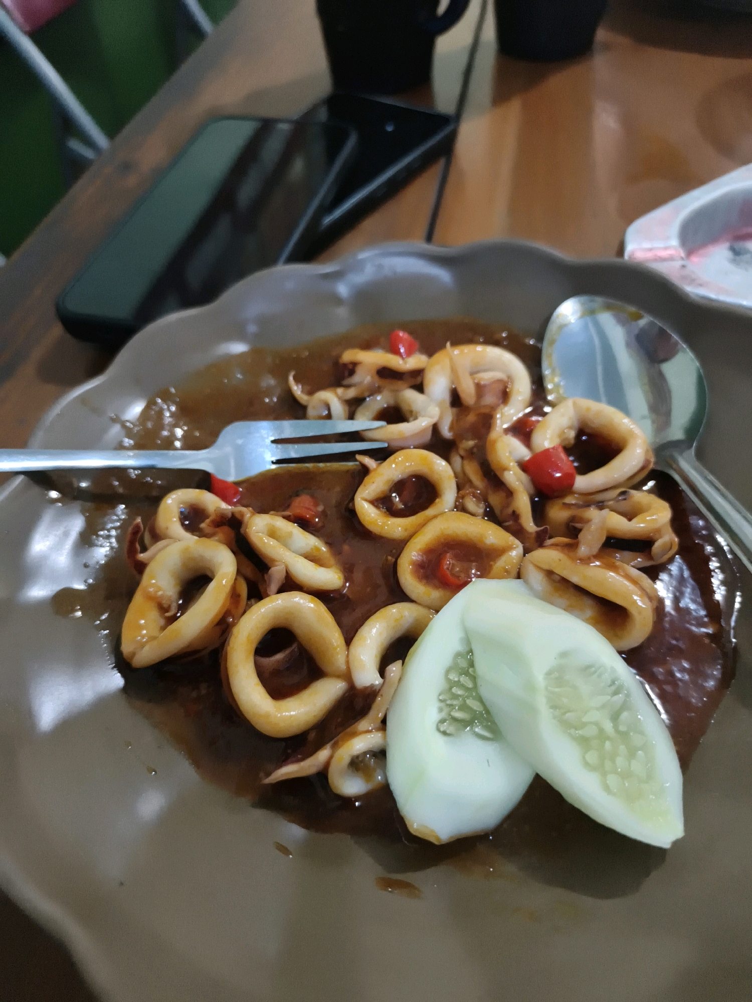 A plate of stir-fried squid is presented on a wooden table, accompanied by two slices of cucumber. The squid rings are coated in a dark, glossy sauce and garnished with small pieces of red chili. A fork rests on the left side of the plate, and a spoon is positioned on the right. In the background, a mobile phone is visible, along with a blurred out-of-focus ashtray and possibly a cup. The overall scene suggests a casual dining setting, likely in Ketapang, Indonesia, where such dishes are commonly enjoyed.