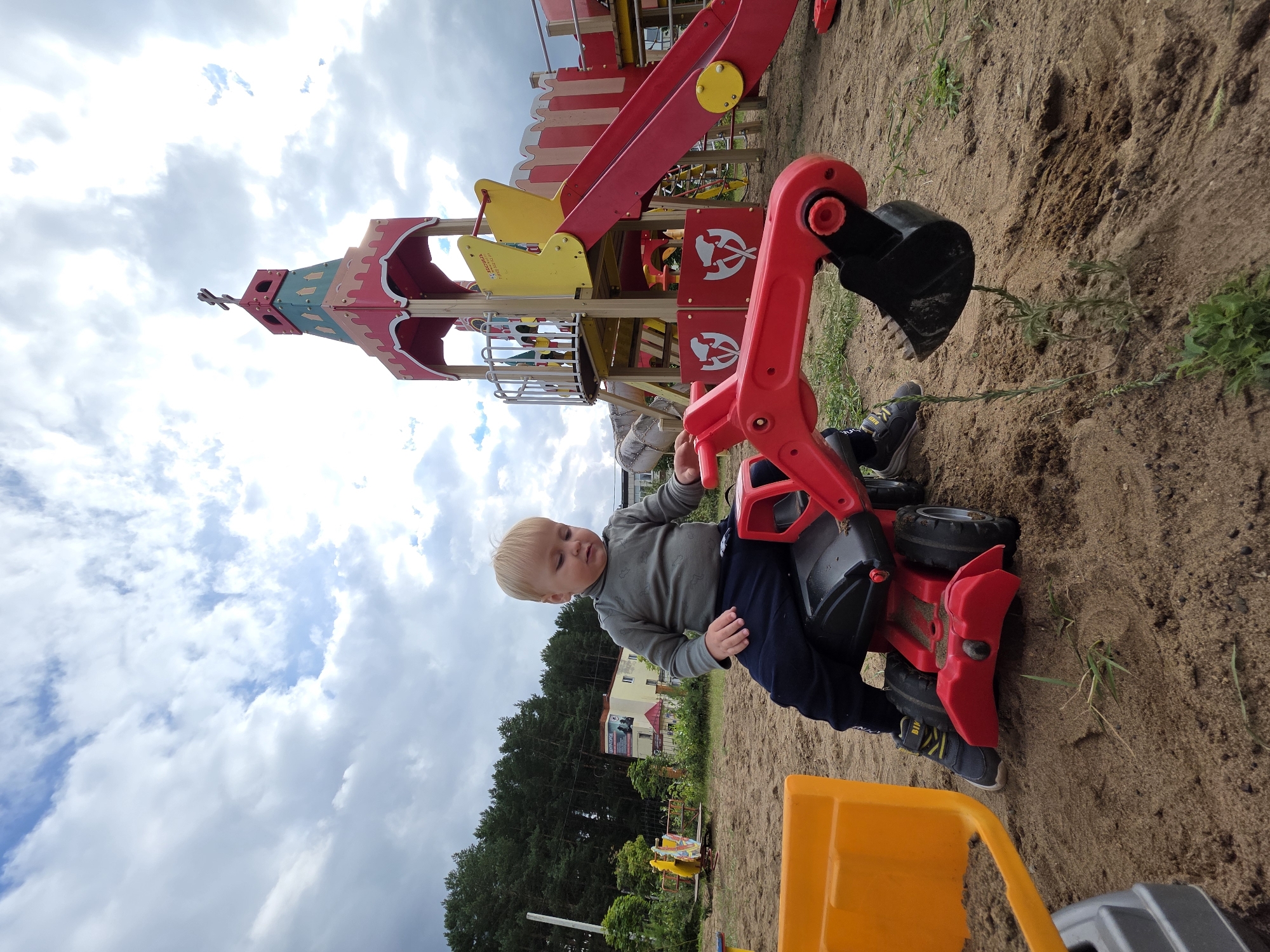 A young Caucasian boy with blonde hair sits on a red toy excavator in a sandy playground. The excavator is positioned in the foreground, with its shovel lowered. The boy is wearing a grey long-sleeved shirt and dark pants. In the background, a colorful playground structure with slides and towers is visible, along with trees and buildings, suggesting an urban or suburban setting. The sky is cloudy with patches of blue. The overall scene depicts a child playing outdoors in a playground. Based on the visible signage in the background that appears to be in Cyrillic script, the location is likely within Russia, potentially in the city of Chaikovsky given the request.