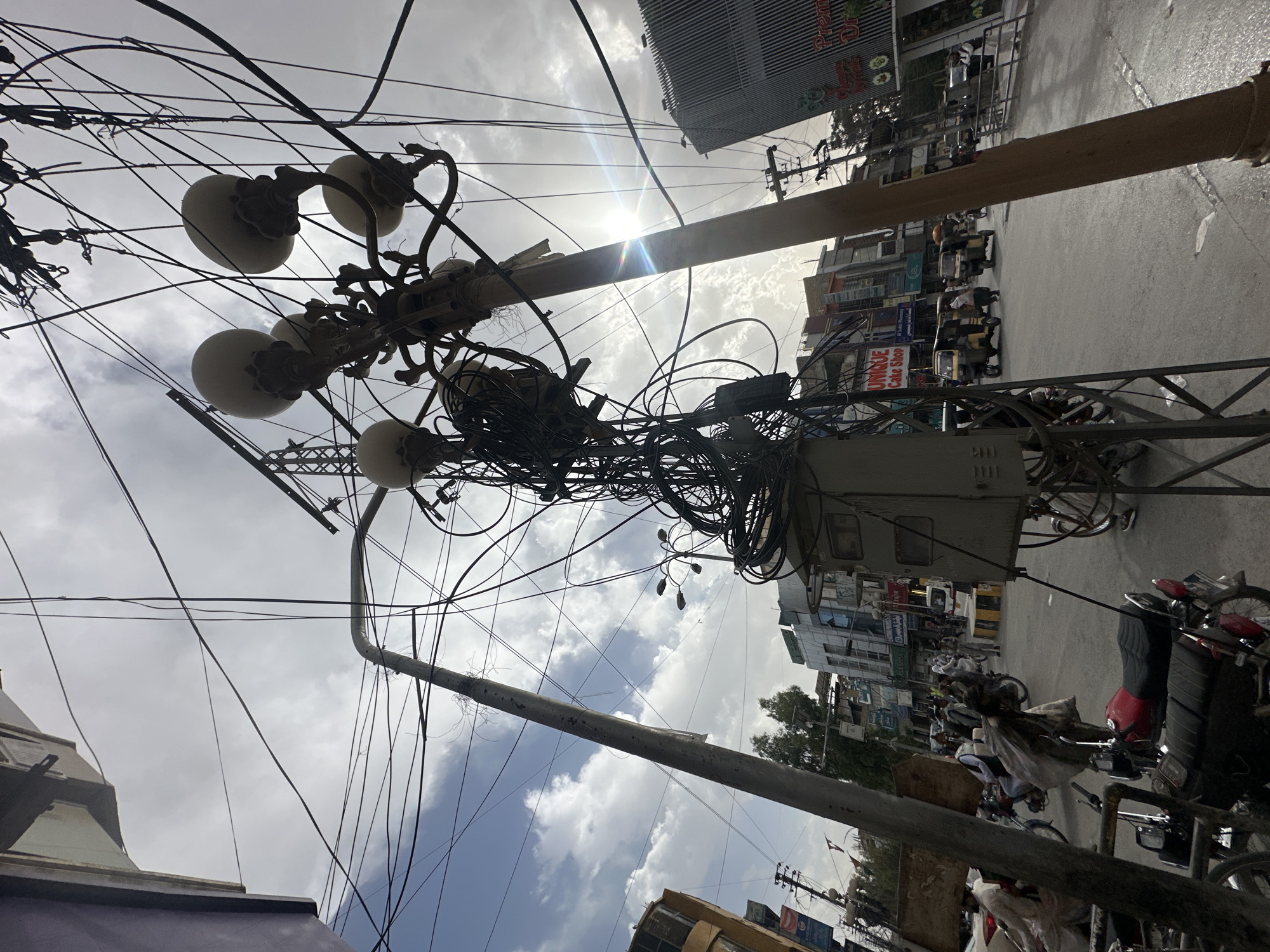 The image shows a street in Quetta City Tehsil, Pakistan. Overhead, a tangle of electrical wires and cables are attached to a utility pole, which also supports a cluster of globe-shaped streetlights. A gray electrical box is mounted on the pole as well. The street scene includes parked motorbikes, auto-rickshaws, and pedestrians. Buildings with various signs and advertisements line the street. The sky is overcast, with the sun partially obscured by clouds.