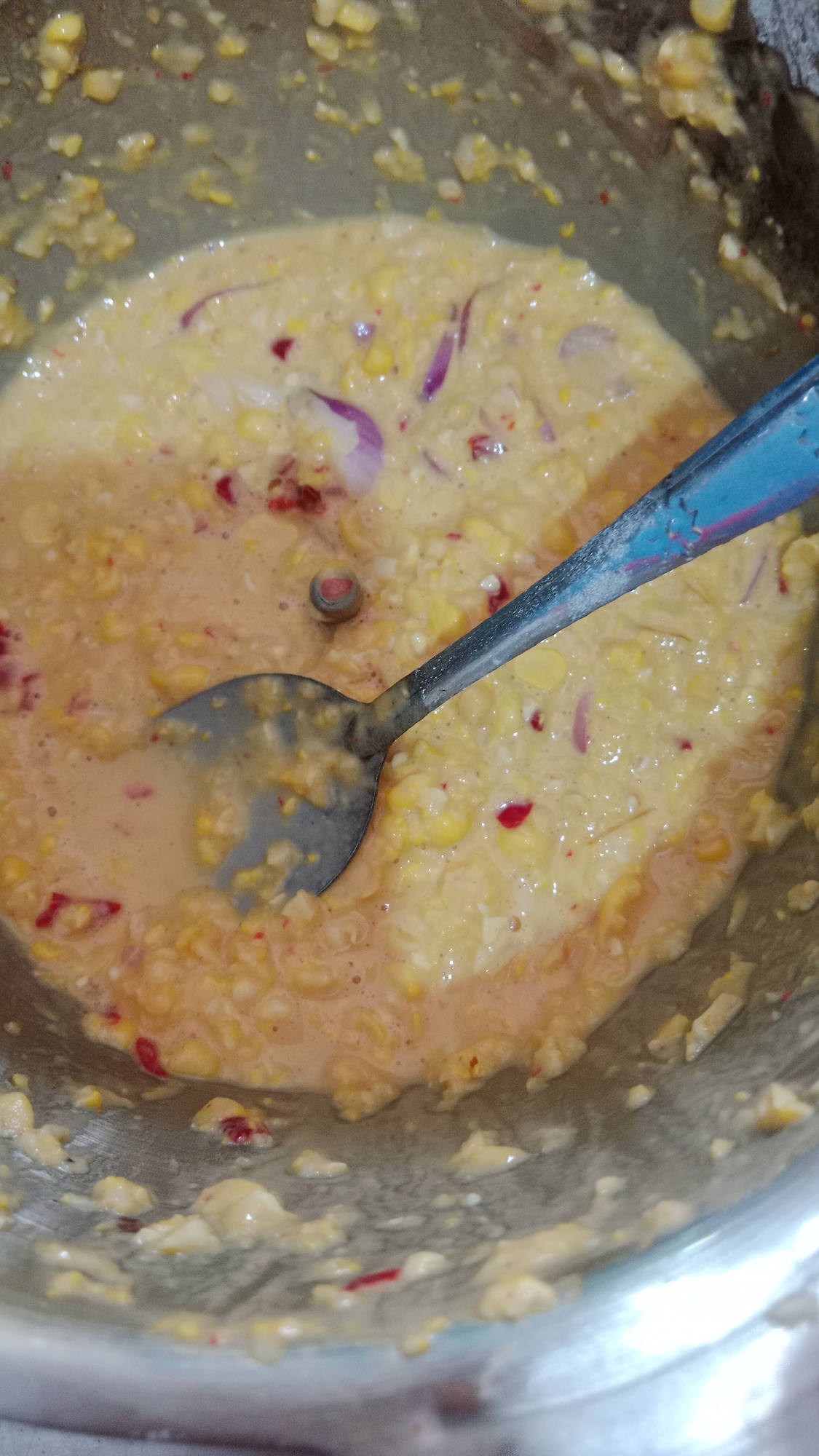 A close-up, overhead shot shows a metal bowl filled with a creamy, yellow mixture containing corn kernels, chopped red onions, and small red flecks, possibly chili. A silver spoon with a blue handle rests in the mixture, with its bowl partially submerged. The mixture appears to be a batter or a type of corn fritter dough. The lighting is somewhat dim, with reflections on the metal bowl. The context of Denpasar, Indonesia, is not visually apparent from the image itself.