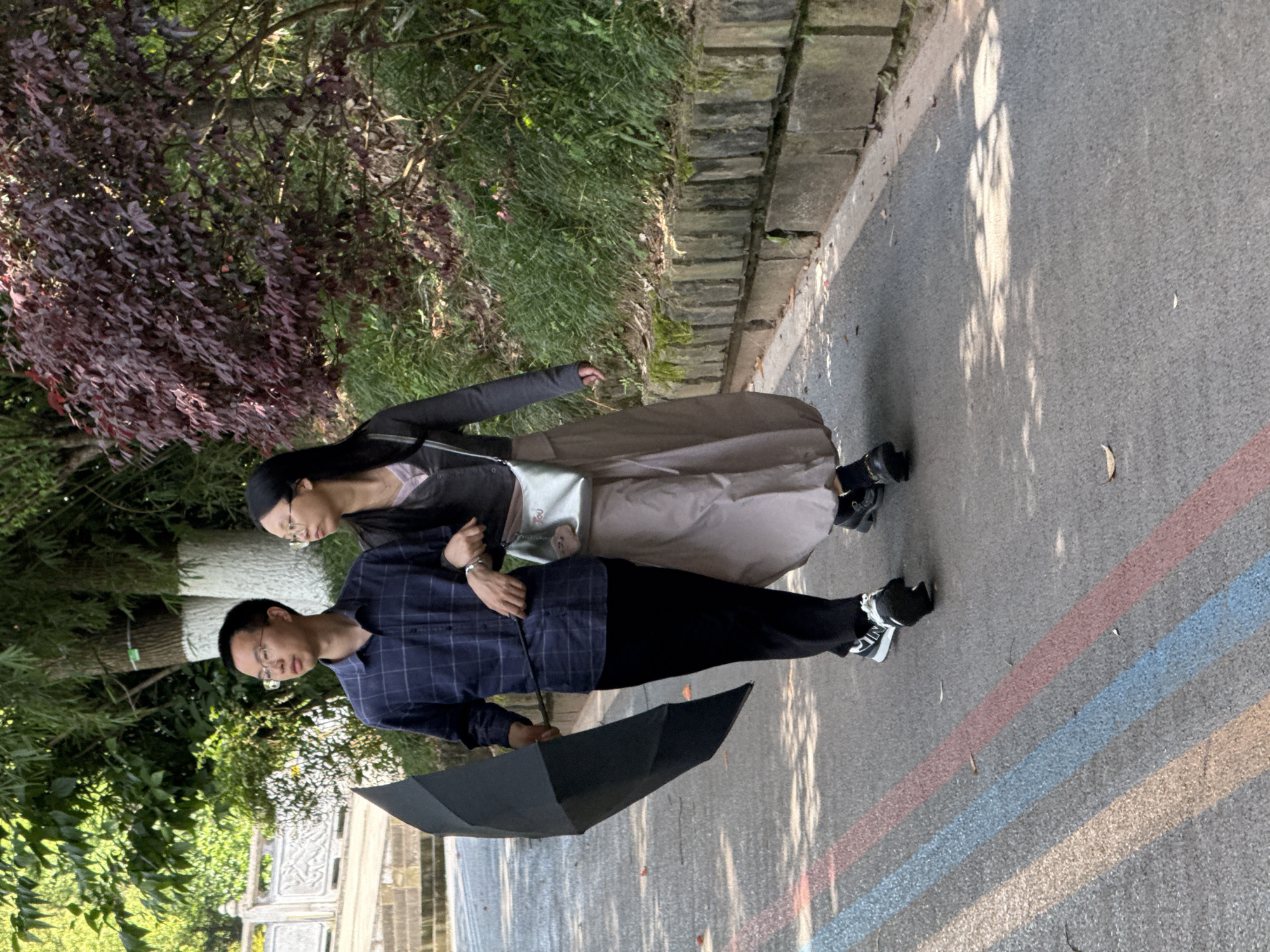 A man and woman are walking side-by-side on a paved walkway in Jiulongpo District, China. The man holds a black umbrella. The woman is wearing glasses, and has a silver purse. A painted rainbow runs diagonally across the walkway. Vegetation and a stone wall are visible on the left side of the frame.