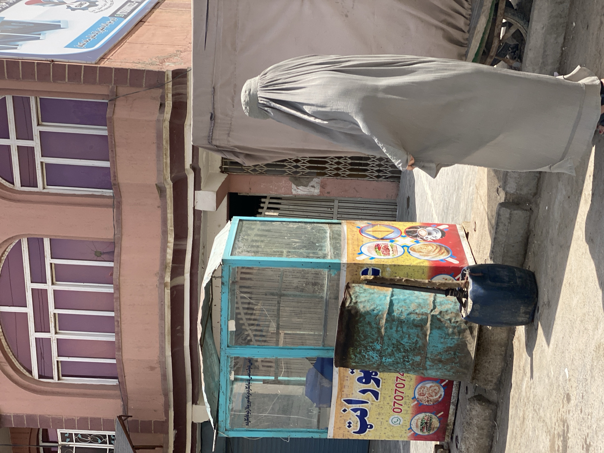 The image depicts a street scene in Kandahar, Afghanistan, likely during daylight. In the foreground, a person wearing a full-length grey burqa, which covers their entire body and face, walks away from the camera. Their right hand is visible, holding the fabric of the burqa. To the left of the person is a colorful food stall with a turquoise-colored frame and glass panels. The stall features advertisements for food items, including burgers and pizzas, and a phone number displayed in yellow text. A large, weathered, teal-colored barrel stands next to the stall. In the background, a multi-story building with arched windows and pinkish-brown stucco walls is visible. The ground is paved with concrete. A large, light-brown tarp covers an object behind the burqa-clad person, partially obscuring a wheeled cart. The overall atmosphere suggests a typical urban setting in Kandahar.