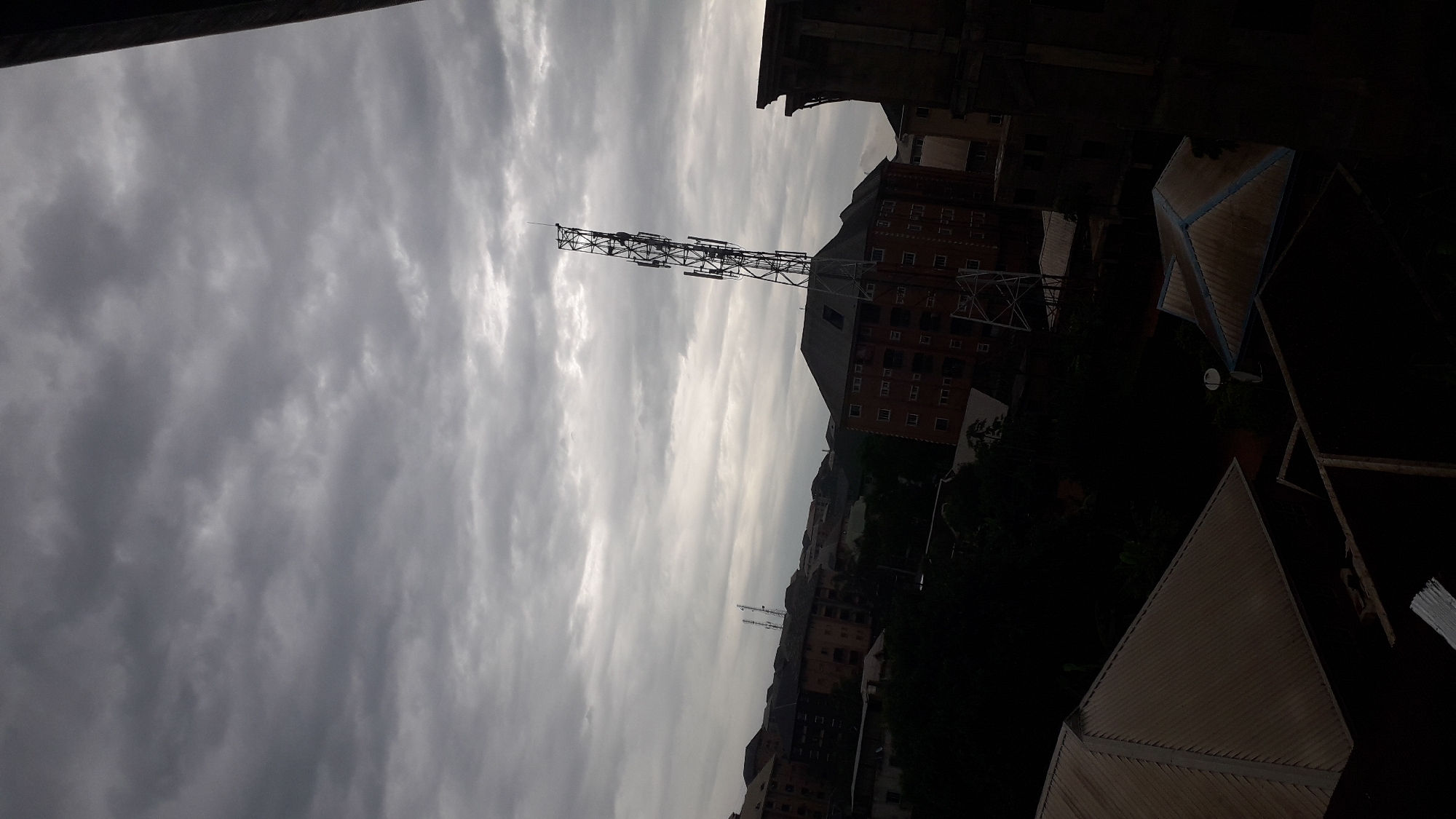 The image shows an overcast sky over a city, with a view of buildings and a cell tower. The foreground features several dark rooftops, one with a peaked, metallic surface and blue trim. In the mid-ground, there are multiple apartment buildings with visible windows and balconies. A prominent cell tower, with its intricate metal structure and multiple antenna arrays, stands tall against the sky. The surrounding environment appears to be urban, with hints of trees and other structures interspersed between the buildings. The lighting suggests it is daytime, but the heavy cloud cover creates a dim, moody atmosphere. The location is identified as Nkpor, Nigeria.