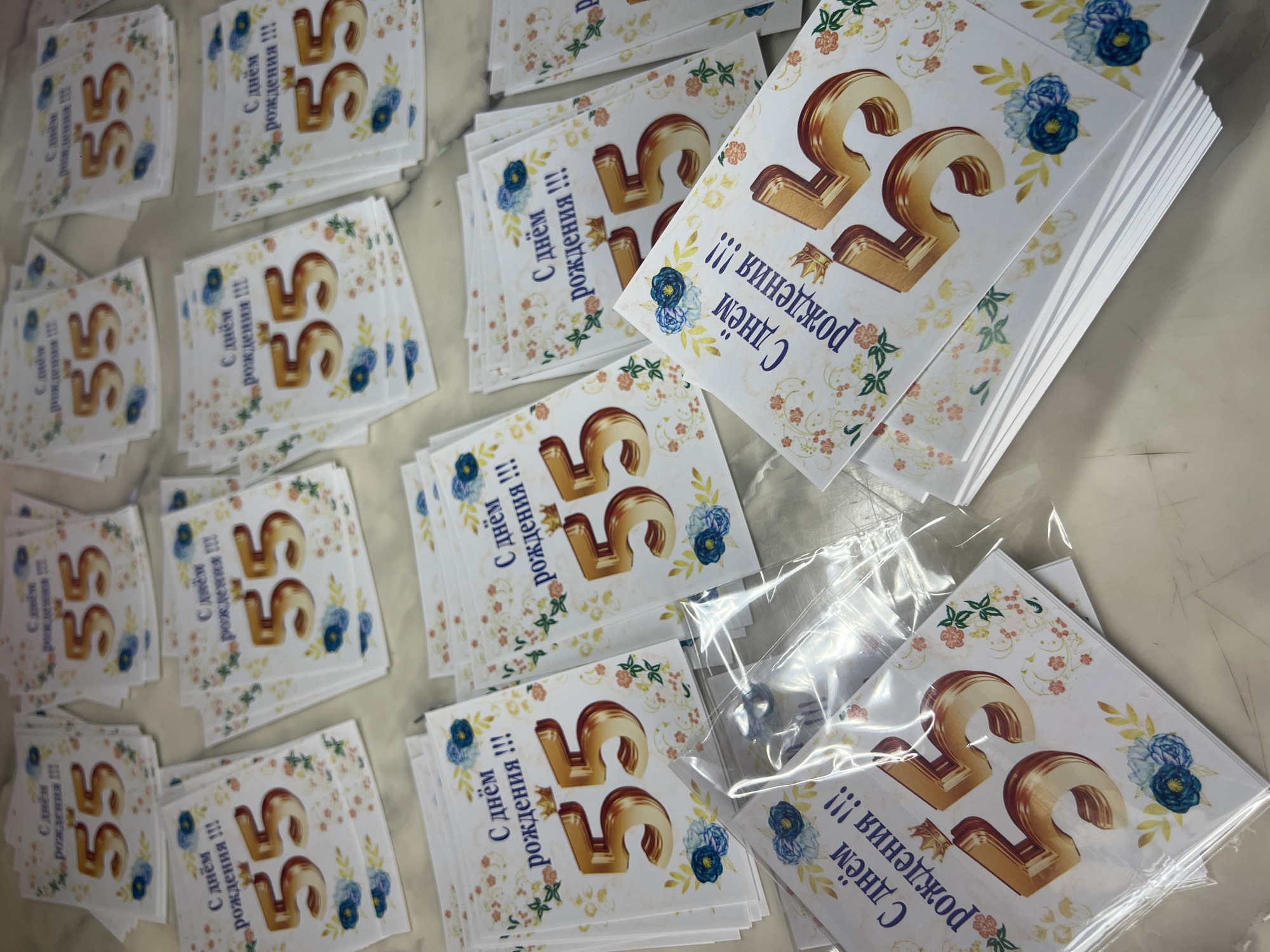 The image shows a pile of greeting cards with the number "55" prominently displayed in gold. The cards also feature floral decorations in shades of blue, orange, and yellow. Some of the cards are individually wrapped in clear plastic. The scene appears to be an indoor setting, possibly a store or a workshop, with a plain, light-colored surface beneath the cards. There are no people visible in the image. The cards are arranged in stacks and some are at an angle, suggesting they are being displayed or prepared for sale. The lighting is consistent throughout the image, indicating a well-lit environment.