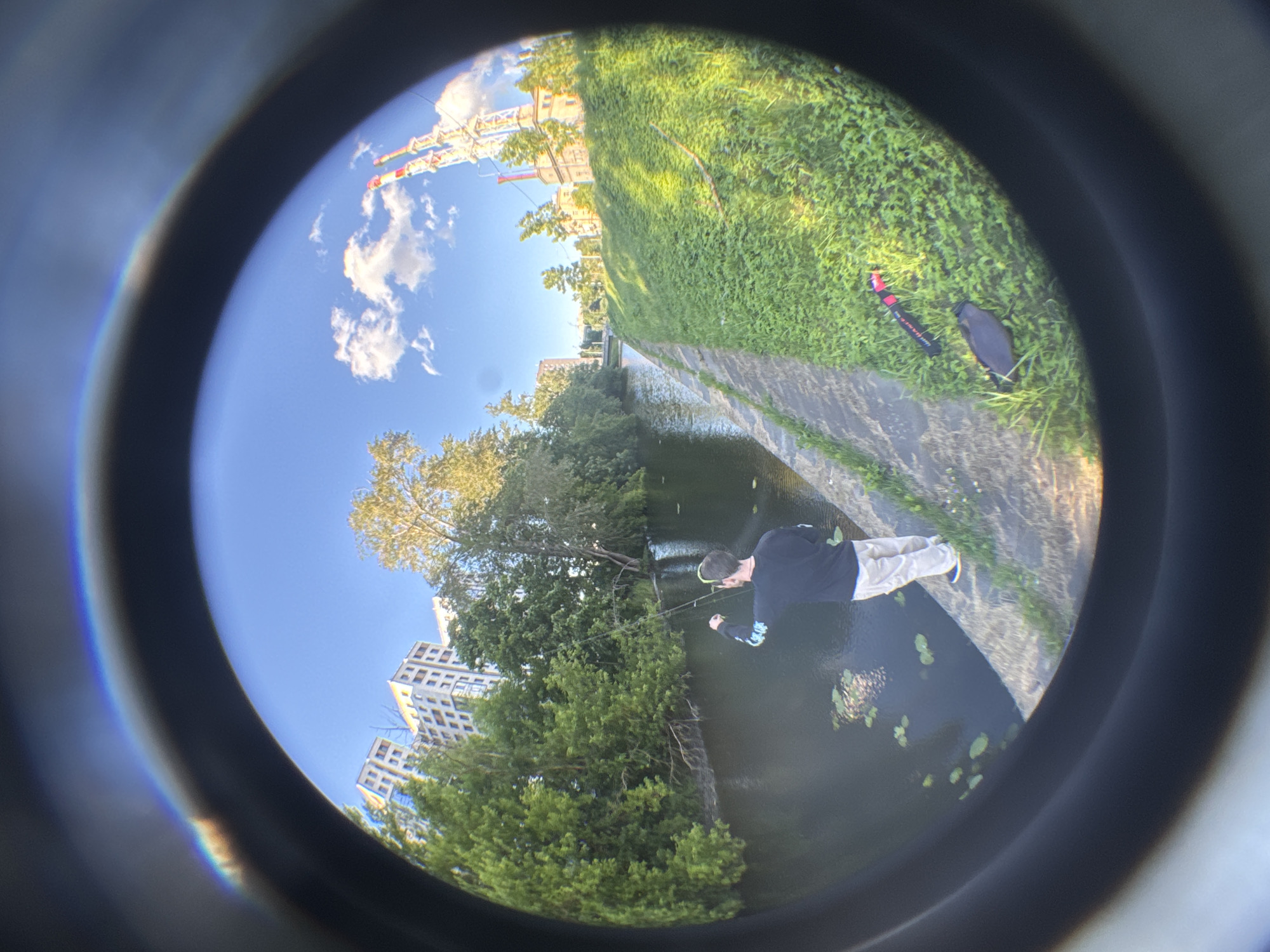 A person is fishing in a canal in Saint Petersburg, Russia. The canal is lined with a concrete embankment and a grassy slope. In the background, there are trees, buildings, and a colorful amusement park structure. The image is taken with a fisheye lens, creating a distorted, circular view with a dark vignette around the edges.