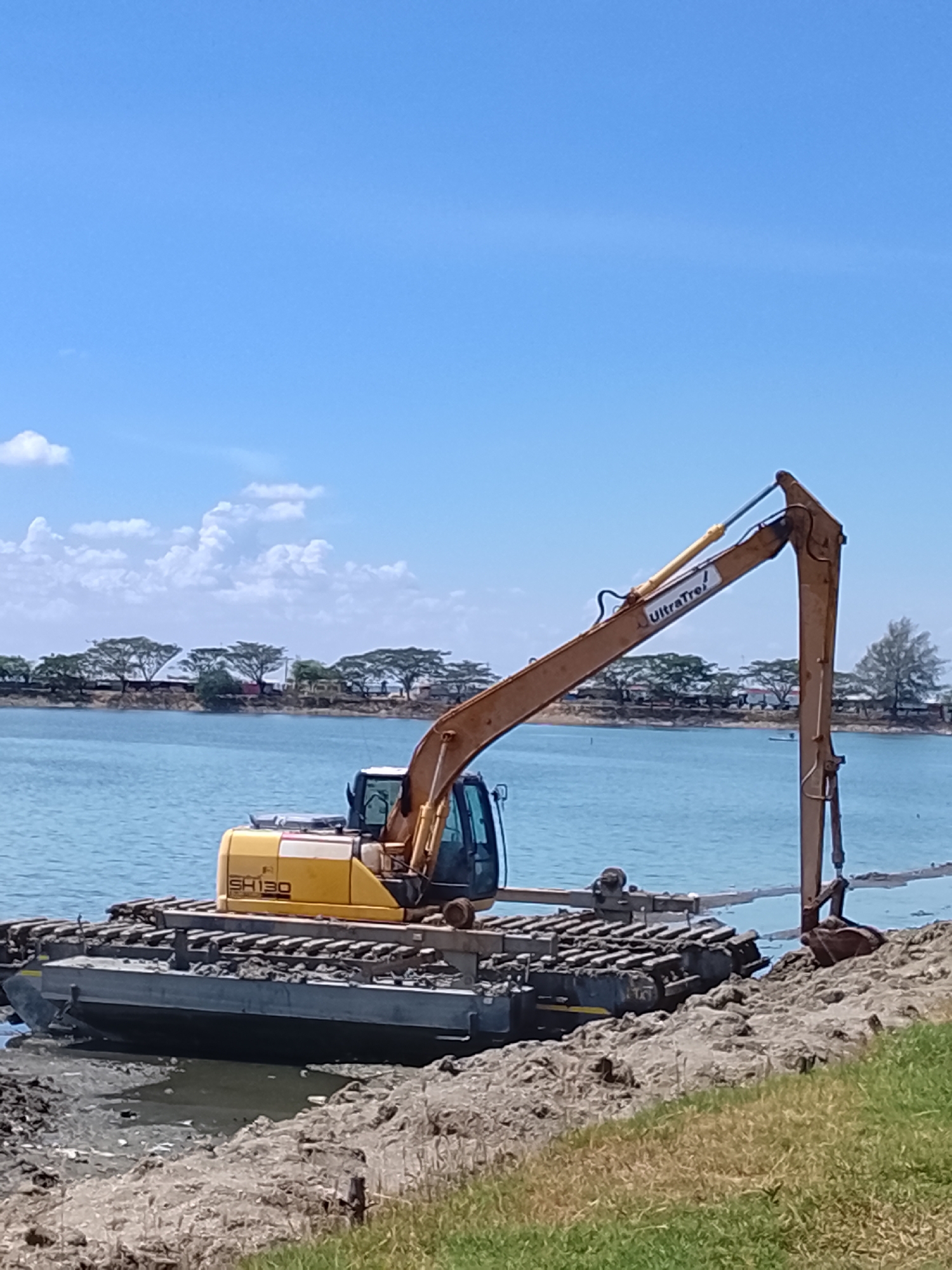 An amphibious excavator, yellow and orange, with the logo "UltraTrec" visible on its boom, is positioned on the muddy shore of a body of water. The excavator is mounted on a pontoon-like base with tracks. Its arm is extended over the water, with a digging bucket at the end. The background features a calm, blue body of water, a tree line along the opposite shore, and a bright blue sky with scattered white clouds. The scene appears to be in Lhokseumawe, Indonesia, a city known for its coastal location and industrial activities. The overall impression is one of ongoing construction or land reclamation work in a tropical environment. There are no people visible in the image.