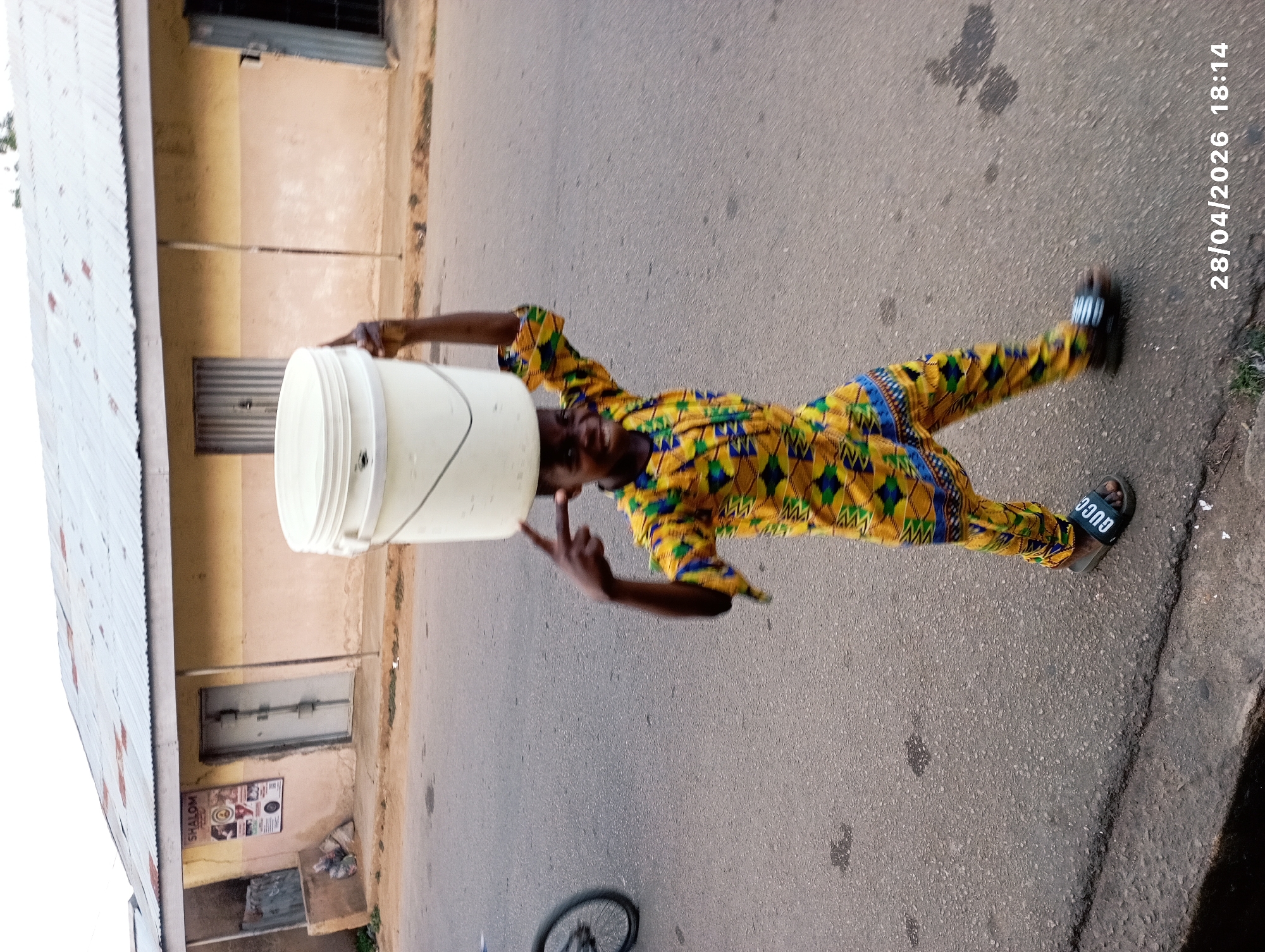 An image taken on April 28, 2026, at 18:14, captures a person skillfully balancing a large white plastic bucket on their head. The individual, appearing male, stands in the middle of a paved road or street, posing confidently. They are dressed in a vibrant, multi-colored two-piece outfit featuring yellow, blue, and green geometric patterns. One hand is raised, making a 'peace' or 'V' sign, while the other arm is bent. The person is wearing black slide sandals, one of which clearly displays the brand name "GUCCI" in white lettering.
The background on the left features a building with a corrugated metal roof, angled upwards towards the sky. On the right, another building with light brown or tan walls is visible, featuring several closed windows or doors. Below one of these, there's a visible sign with the word "SHALOM" and what appears to be a promotional poster or menu board. The road surface is a dark grey asphalt with some minor debris. A portion of a bicycle wheel can be seen on the lower left side of the frame. The lighting suggests early evening, consistent with the timestamp. The overall scene reflects daily life in an urban or semi-urban setting, likely in Jos, Nigeria, given the provided context.