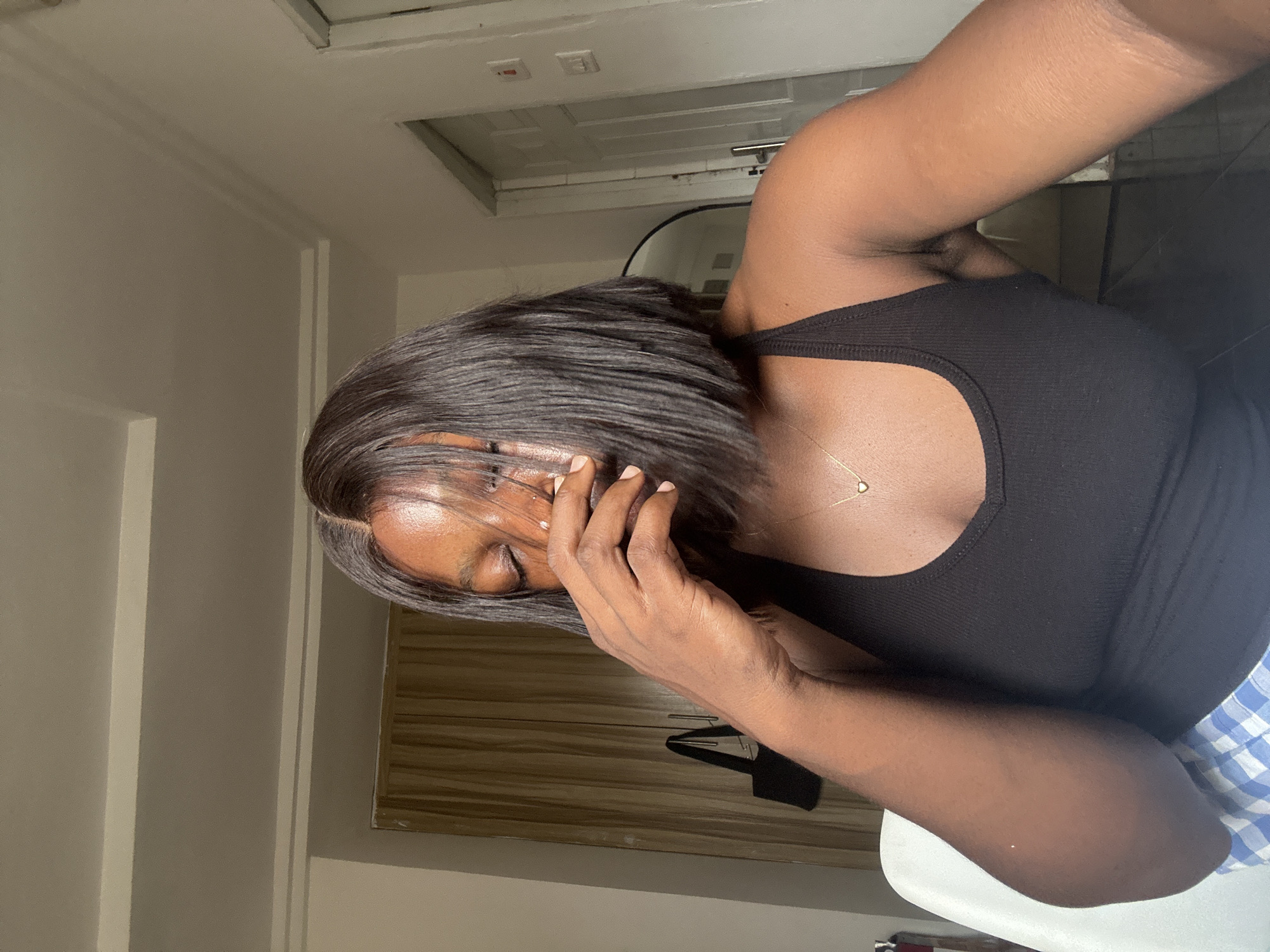 A dark-skinned woman with short, dark hair is indoors, obscuring her face with her hand. She is wearing a black tank top and a thin gold necklace. The background includes a white door, a white ceiling with recessed lighting, light-colored walls, and a wooden cabinet. The presence of modern furniture and design elements suggests an urban setting in Abuja, Nigeria. There is a plaid fabric visible to the right. The image is oriented vertically.
