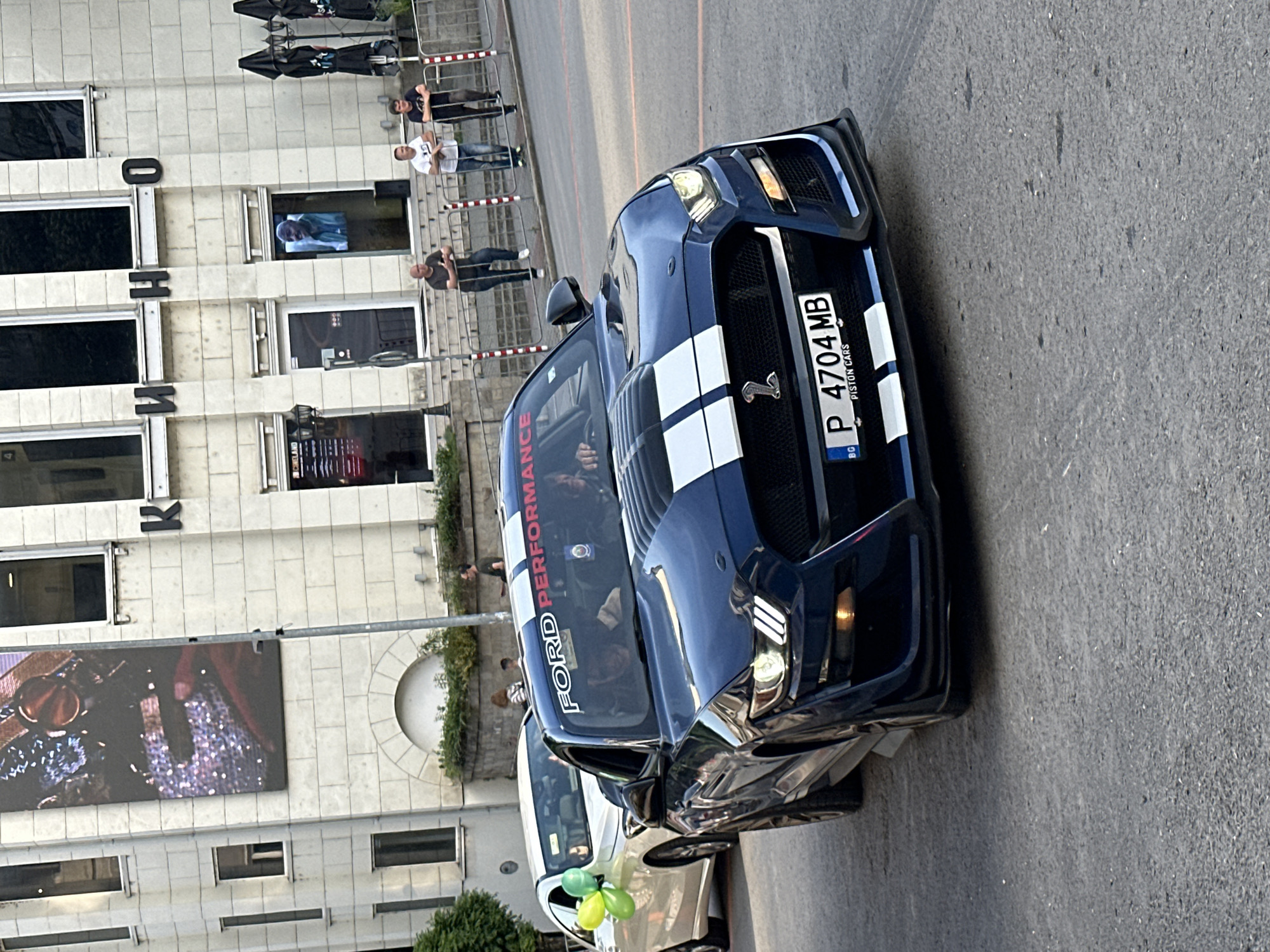 The image shows a blue Ford Mustang with white racing stripes parked on a street in Veliko Tarnovo, Bulgaria. The car has a Bulgarian license plate, P 4704 MB. The words "FORD PERFORMANCE" are visible on the windshield. The building in the background has the letters "K" and "O" visible on its facade. There are people walking behind a metal railing in the background. A white car with green and yellow balloons is parked behind the Mustang. The road is asphalt.