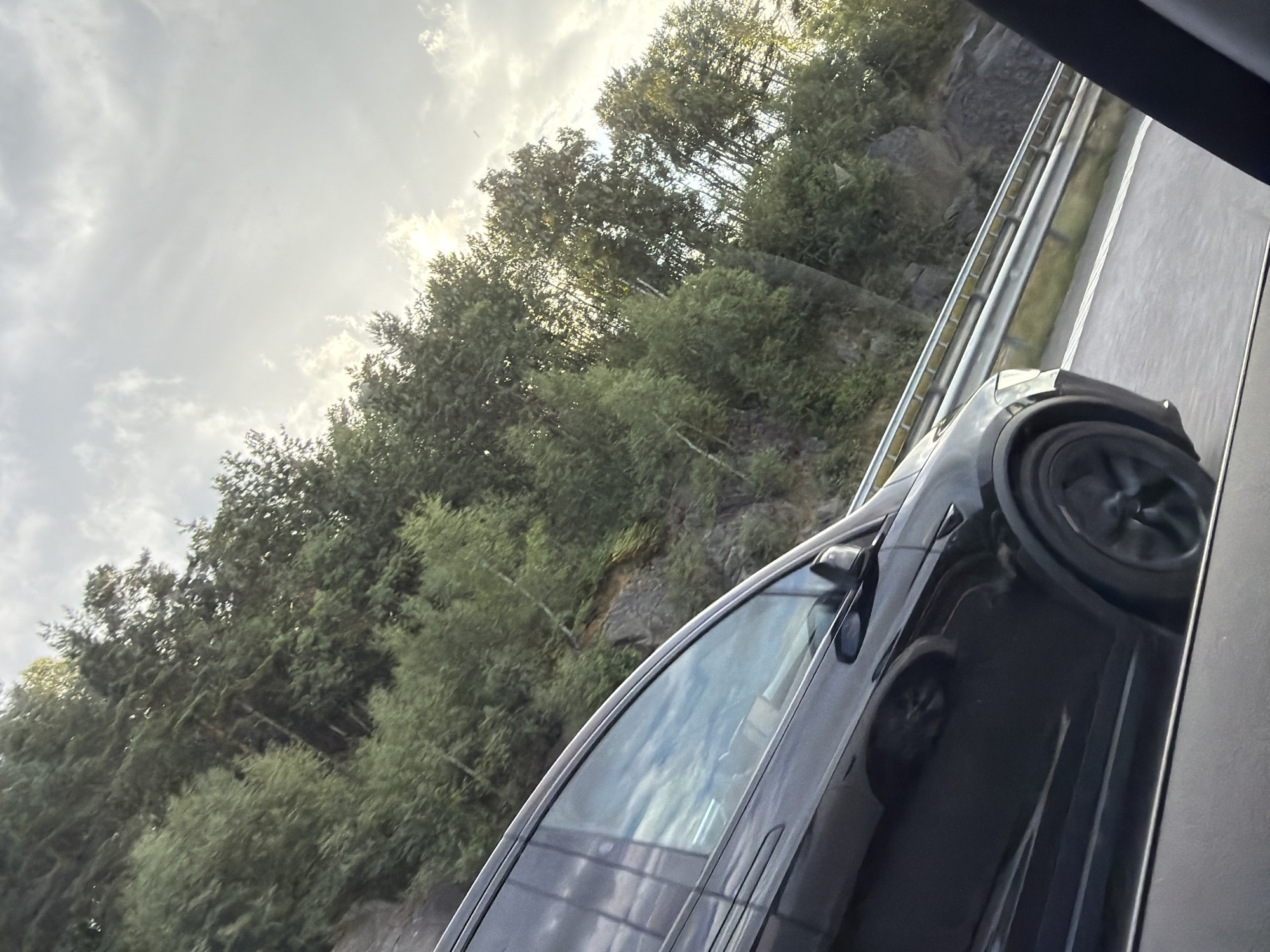 The image, taken from inside a vehicle on a sunny day, shows a black Tesla SUV driving on a highway. The car is in motion, with its wheels blurred. The highway has a metal guardrail and white lane markings. To the right of the highway, a rocky hillside covered in dense green trees rises steeply. The sky is partly cloudy, with sunlight breaking through the clouds, casting a bright glow. The overall scene suggests travel through a scenic, possibly mountainous or hilly, natural environment. The location is identified as Åsa, Sweden.