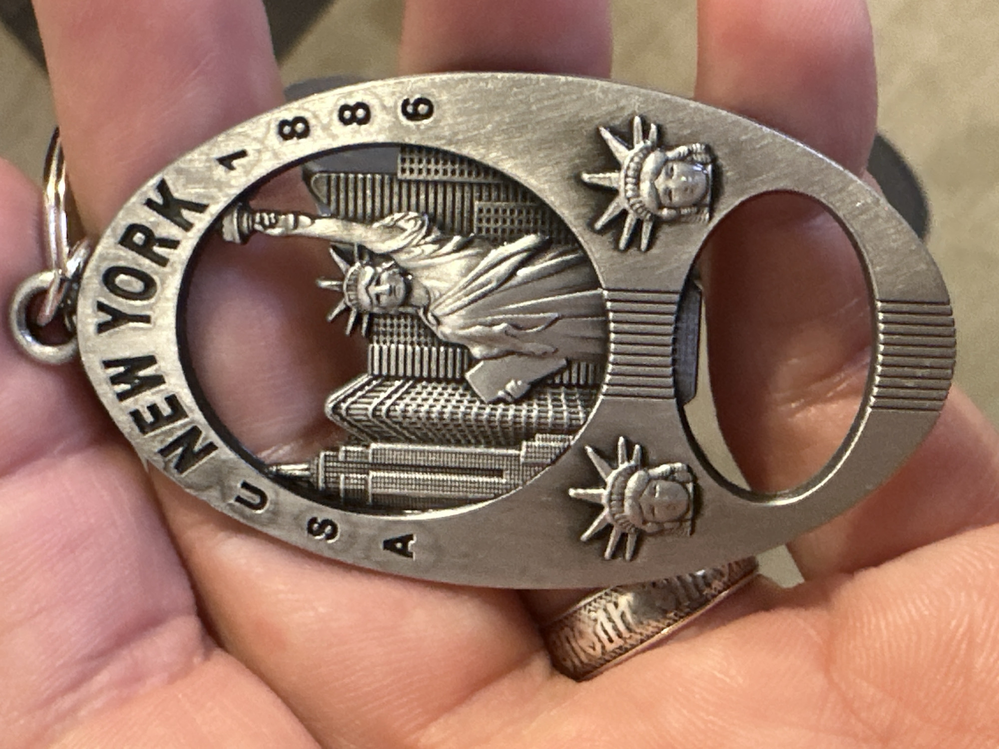 A metal keychain with the inscription "NEW YORK U.S.A. 1886" is held in a person's hand. The keychain features a depiction of the Statue of Liberty and New York City skyline, along with two smaller representations of the Statue of Liberty's head at the bottom. The keychain has a cutout forming a bottle opener shape. The background is a hand and a textured surface, possibly a table. There is no information in the image to suggest a location in Uzhhorod, Ukraine.