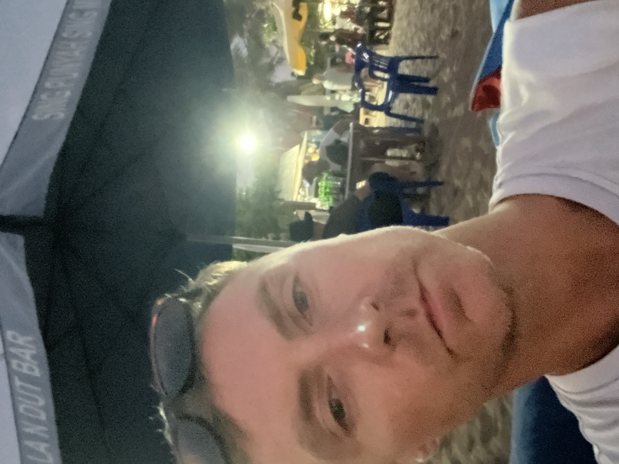 The image is a selfie taken by a man with light skin wearing a white shirt. He has sunglasses on the top of his head. The background appears to be an outdoor dining area at night. In the background, there are people seated at tables with blue plastic chairs. There are some awnings or canopies visible, and lights illuminating the scene. Part of an umbrella with the words "LANDUT BAR" is visible. The picture was likely taken in Kuta, Indonesia.