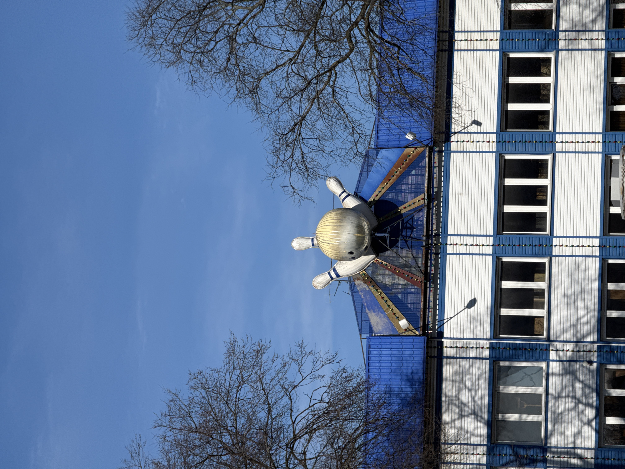 The image shows the roof of a building with a large bowling pin and ball sculpture on top of it. The building has a blue roof and white walls with blue trim and is located in an unknown city. The sky is blue and there are bare trees visible in the foreground.