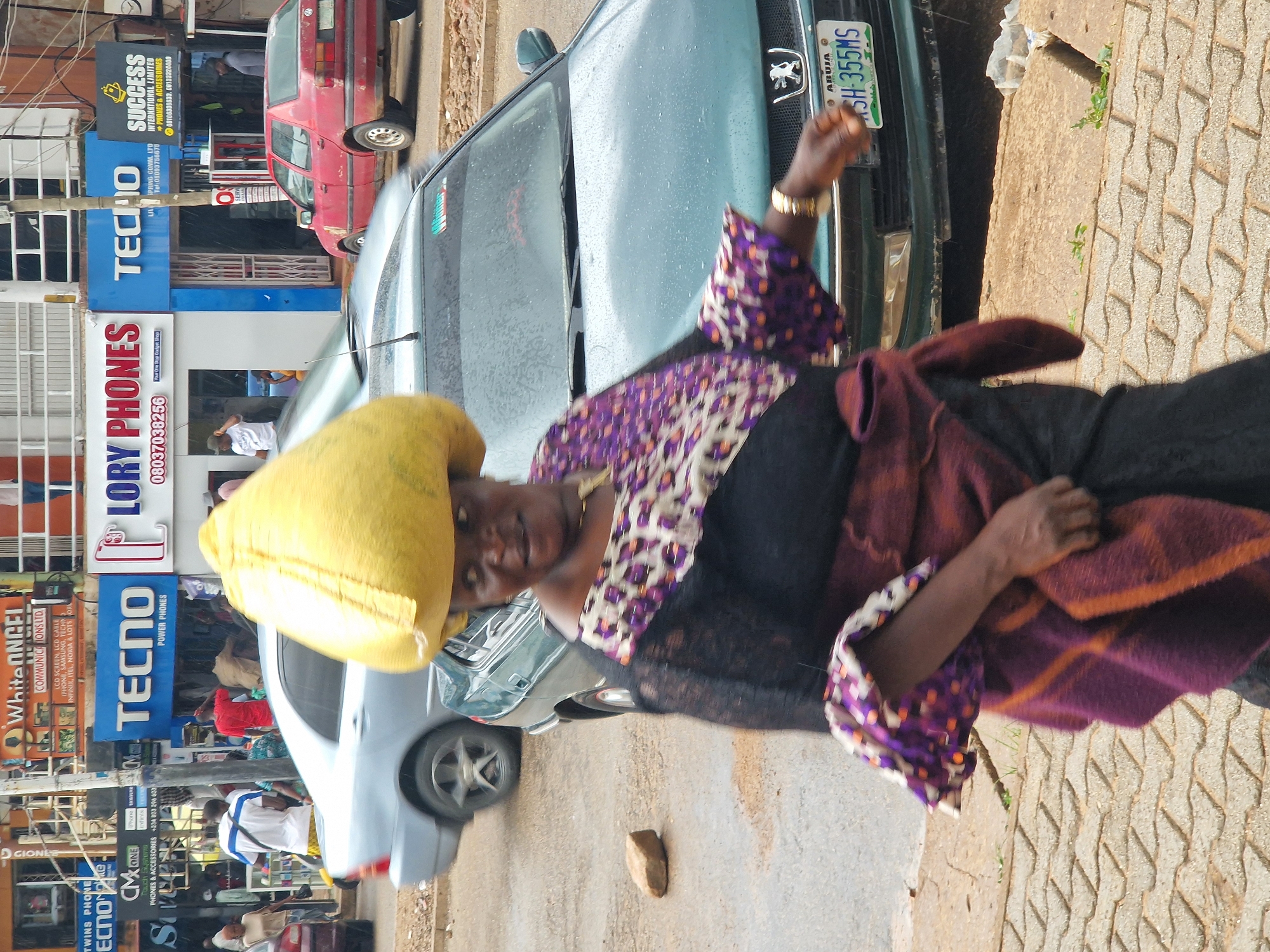 In Jos, Nigeria, a woman with a large yellow head covering walks in front of a green sedan with a license plate reading "SH-355MS." She is wearing a black dress with a patterned purple and white overlay on the sleeves and shoulders, and a reddish-purple sash around her waist. Her right arm is extended as if gesturing or walking. Behind the green car, a red Volkswagen Golf is parked. The background shows a street scene with various shops, including one with a large "TECNO POWER PHONES" sign. People are visible inside and outside the shops. The ground is wet, suggesting it has recently rained.