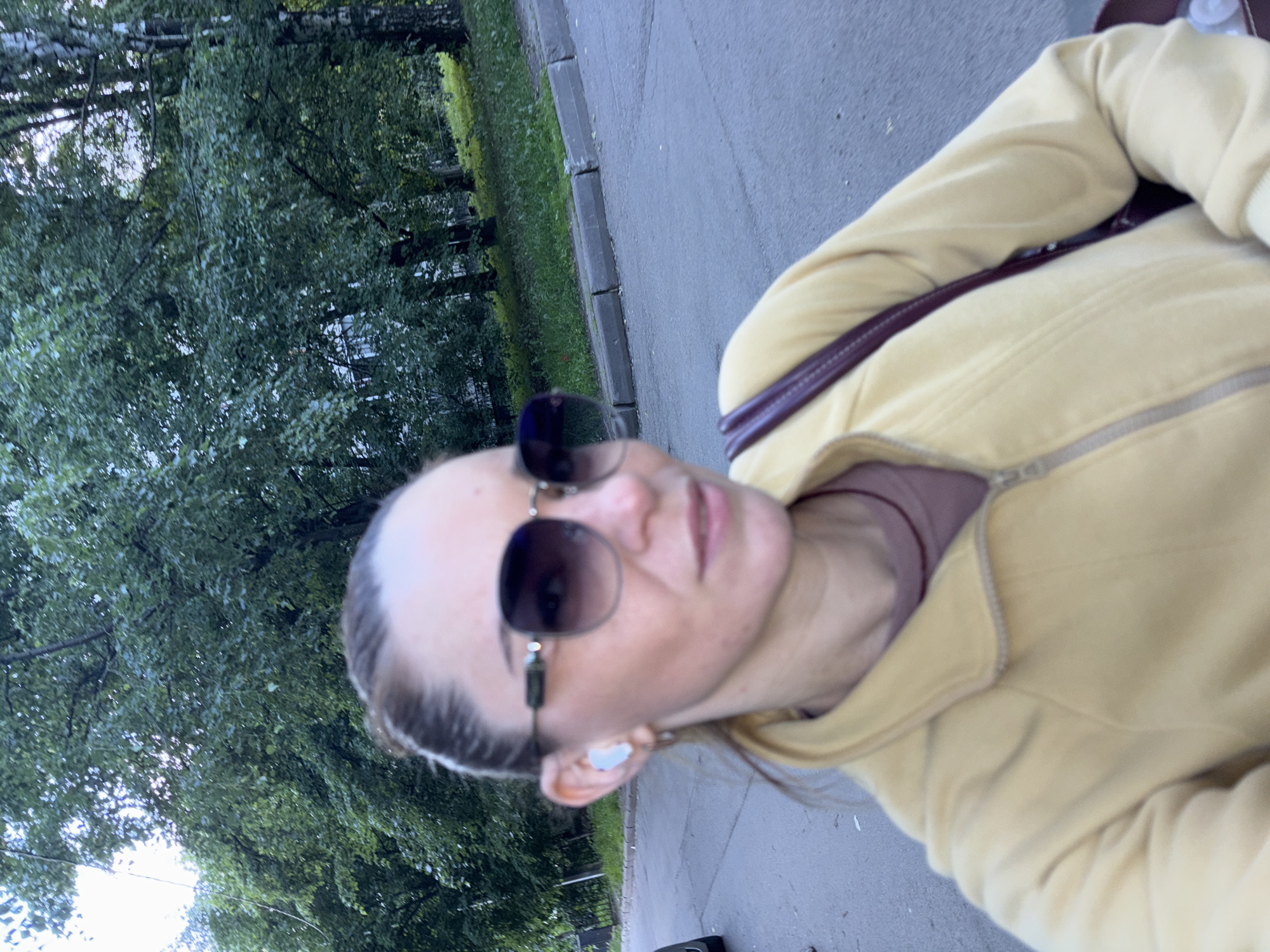 A medium shot captures a woman in sunglasses and a light yellow zip-up jacket walking outdoors. She has her hair pulled back and is wearing a white earbud in her left ear. A dark red strap of a bag is visible over her right shoulder. The background shows a paved area, a sidewalk with a curb, and lush green trees and foliage. The scene appears to be a residential street in Sinyavino, Russia.