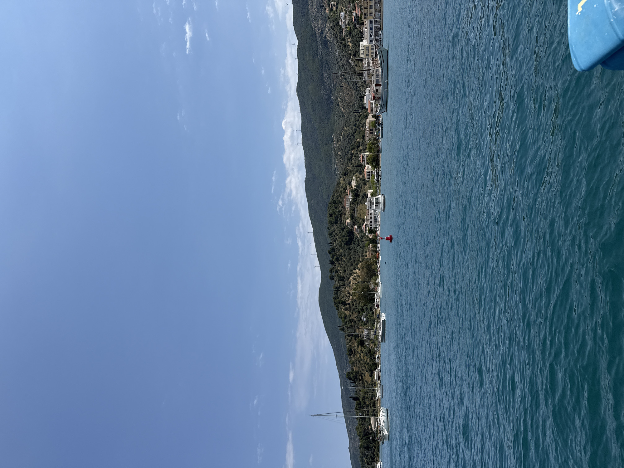 A scenic view captures the city of Poros, Greece. The image focuses on the coastline, where numerous houses and buildings are nestled along the edge of a steep, green-covered hill. The buildings vary in size and architecture. The blue sea dominates the right side of the frame, reflecting the sunlight. Wind turbines are visible atop the hill. The sky above is a clear blue, dotted with a few light clouds. Several sailing boats are docked along the coast. A red buoy floats in the water near the shore. A section of a blue object is on the top right.
