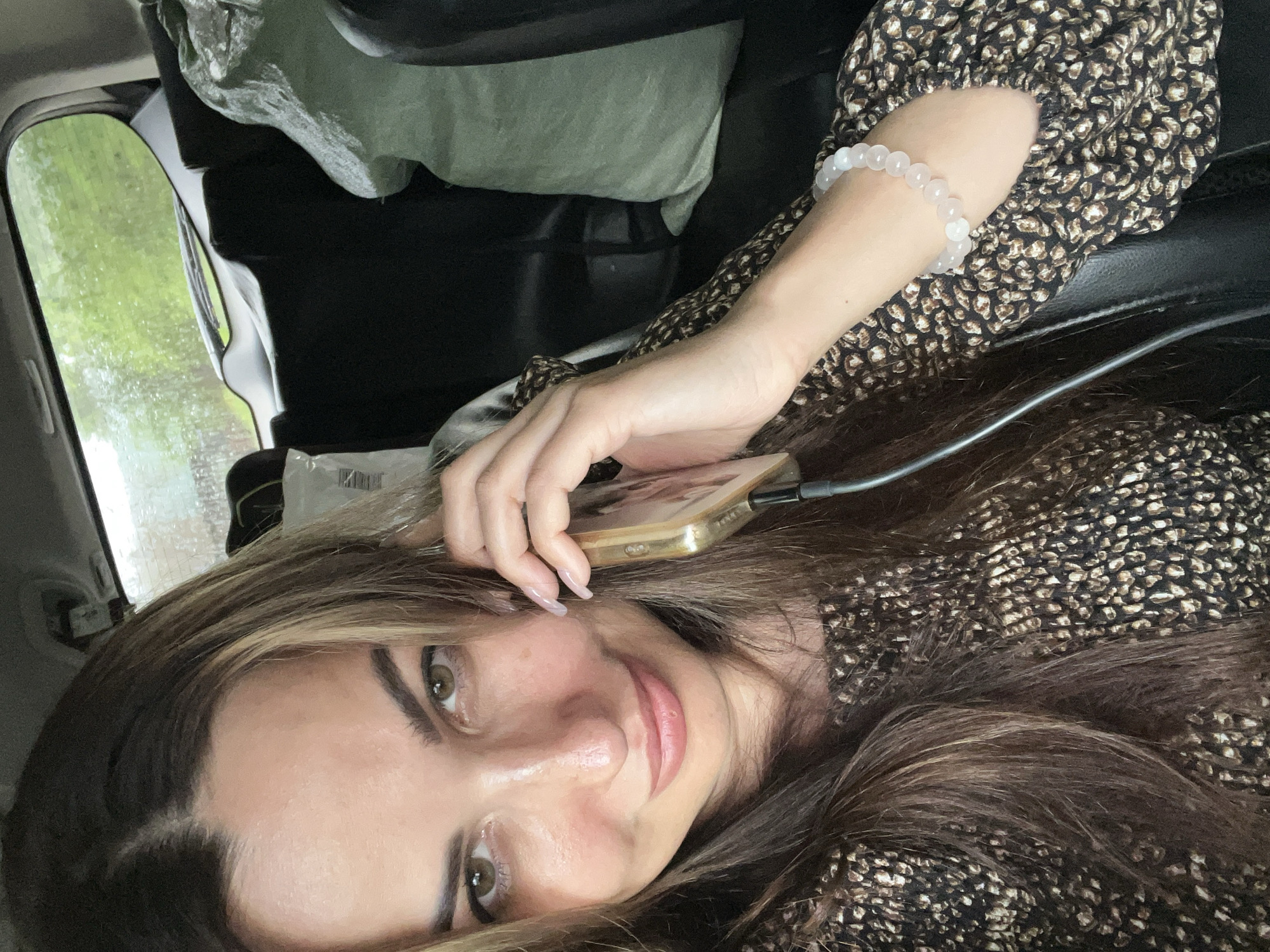 A medium shot shows a woman in the back seat of a car, holding a smartphone to her ear. She is wearing a long-sleeved, patterned top and a white beaded bracelet on her left wrist. Her hair is long and brown, with some lighter streaks. The interior of the car is dark, with black seats. Outside the car window, green trees are visible through the rain. Two large, light green bags are in the back seat behind her. The location is unknown.