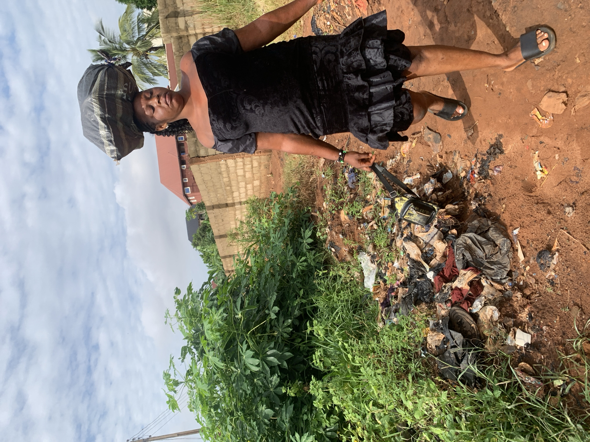 A woman in Nsugbe, Nigeria, is carrying a large bundle on her head. She is wearing a short, ruffled black dress and sandals. A small bag is slung over her shoulder. She stands on a dirt path next to a pile of trash, amidst vegetation. A brick building and palm trees are visible in the background under a cloudy sky.