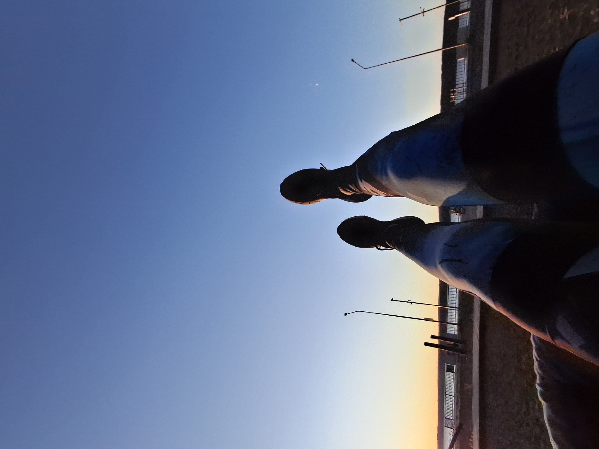 The image shows a low-angle view of a person's legs and feet, wearing dark boots and patterned leggings, against a clear blue sky with a hint of sunset colors near the horizon. The person appears to be sitting or reclining outdoors. In the background, there is a body of water, a shoreline, and a promenade with streetlights and railings. The setting suggests a public space near water, possibly a park or embankment. The lighting indicates it is either sunrise or sunset. The location is in the city of Chaikovsky, Russia.