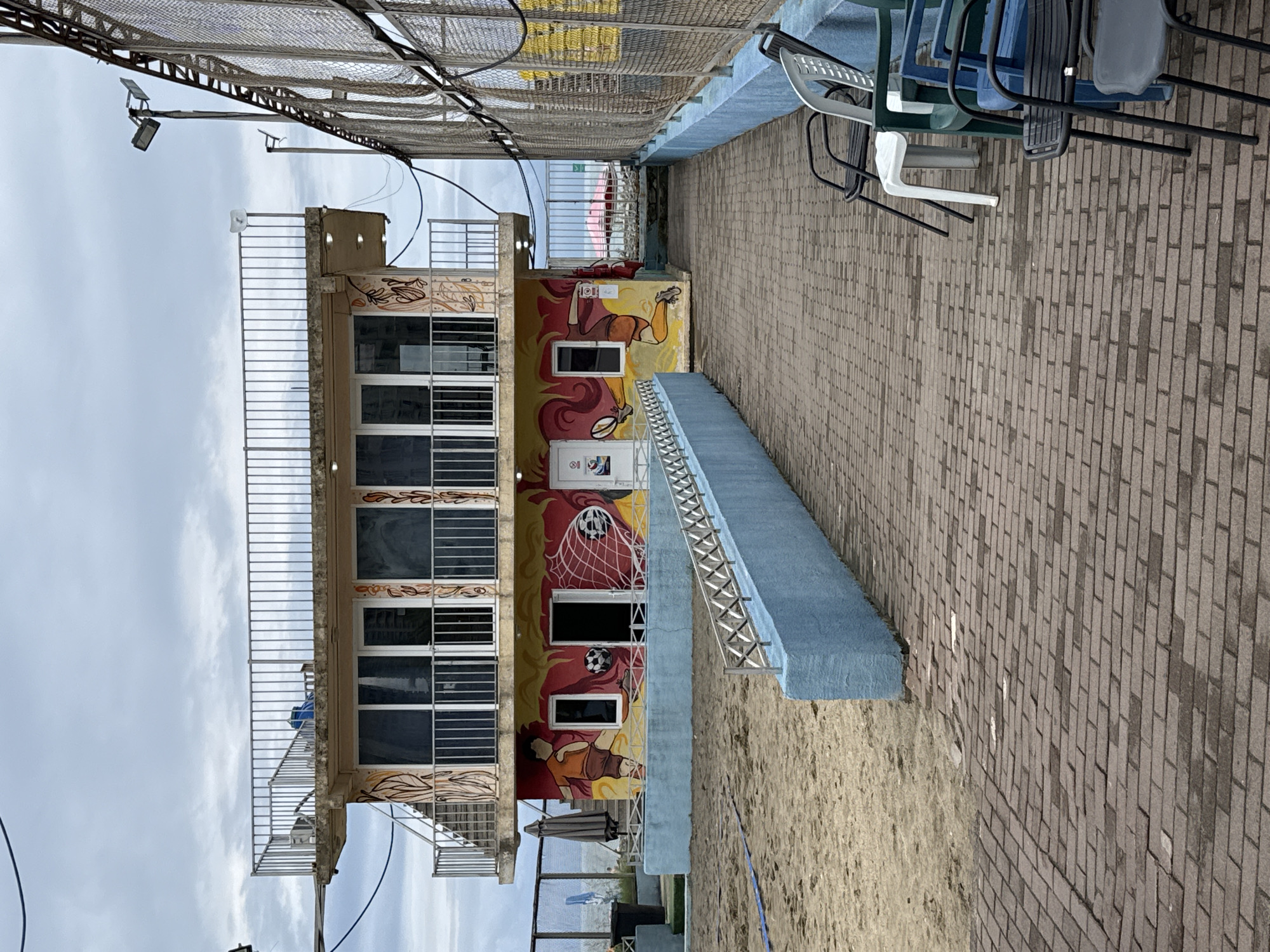 The image depicts a building with two floors, decorated with colorful murals of soccer players and a soccer ball. The building has large windows on both floors and a balcony on the upper level with a metal railing. The ground floor opens onto a sandy area, possibly a beach or sports court. In front of the building, there is a paved walkway with several plastic chairs lined up along the side. To the right, a tall chain-link fence encloses a sports field with floodlights. The sky is overcast. This scene is located at the Batumi Boulevard in Batumi, Georgia, known for its recreational facilities and proximity to the Black Sea.